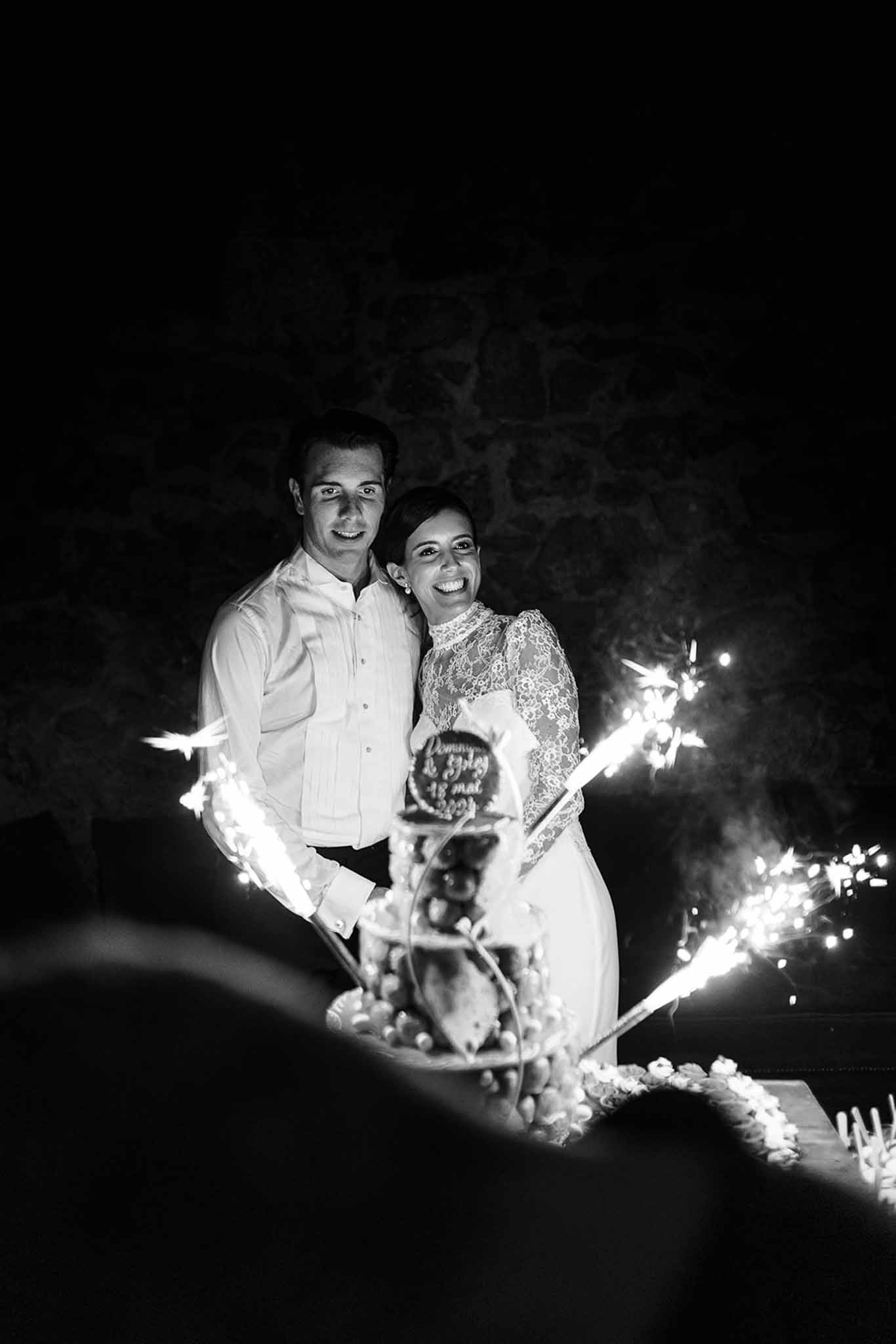 Black and white of couple cutting croquembouche tower surrounded by sparklers against stone wall at night