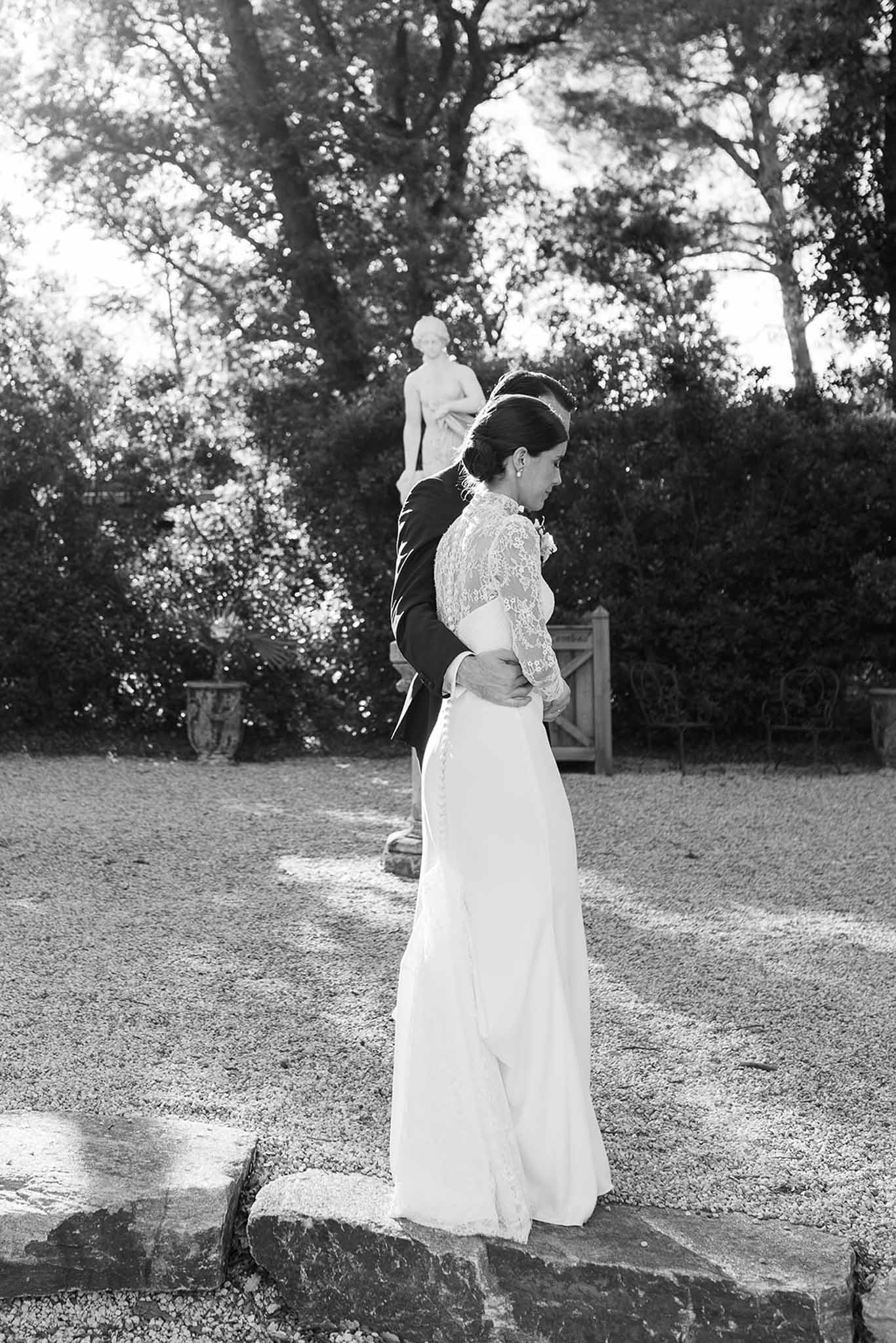 Black-and-white portrait of bride and groom embracing in a formal garden with statue and trimmed hedgerows