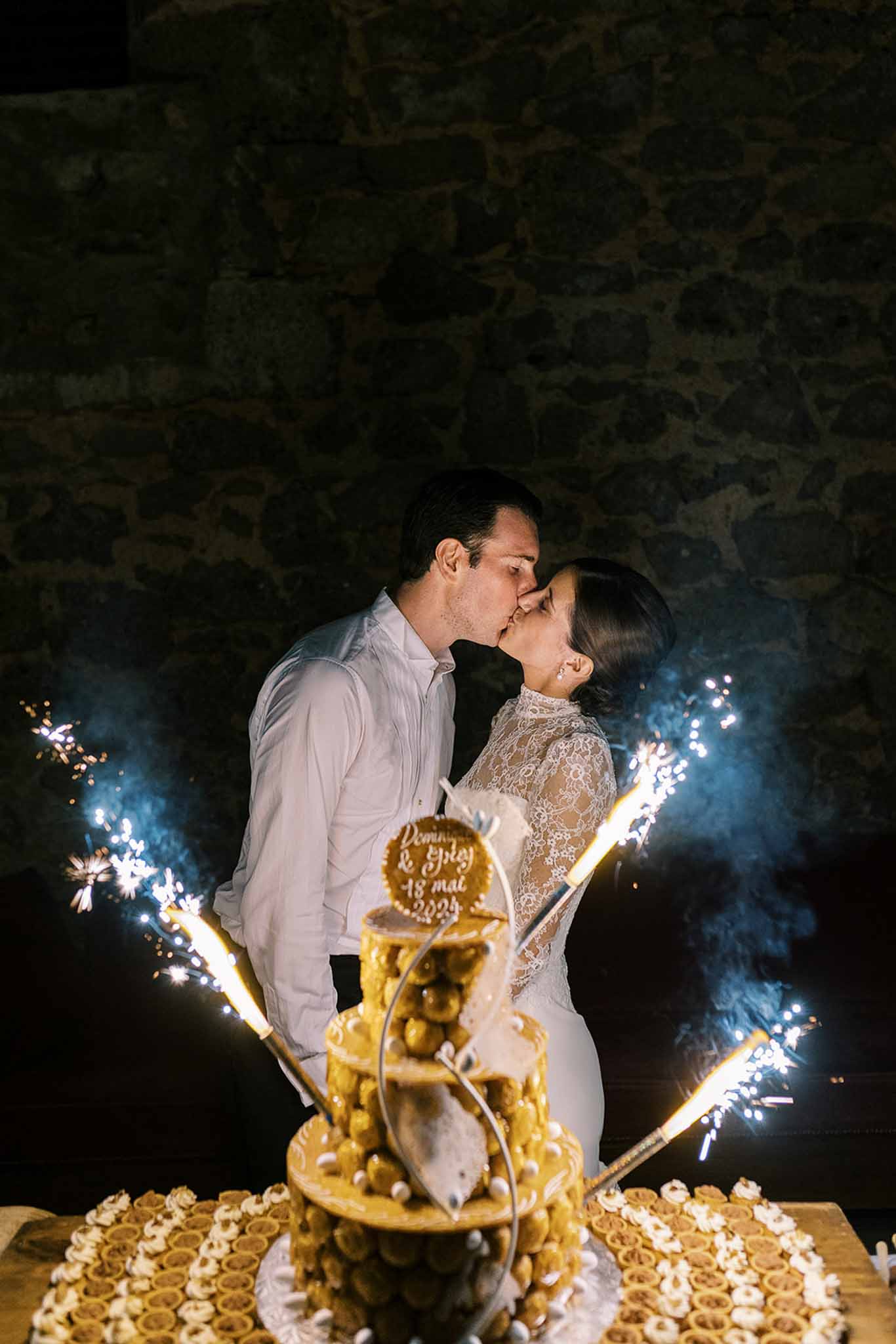 Bride and groom kiss behind croquembouche tower with lit sparklers during evening cake cutting at stone-walled reception
