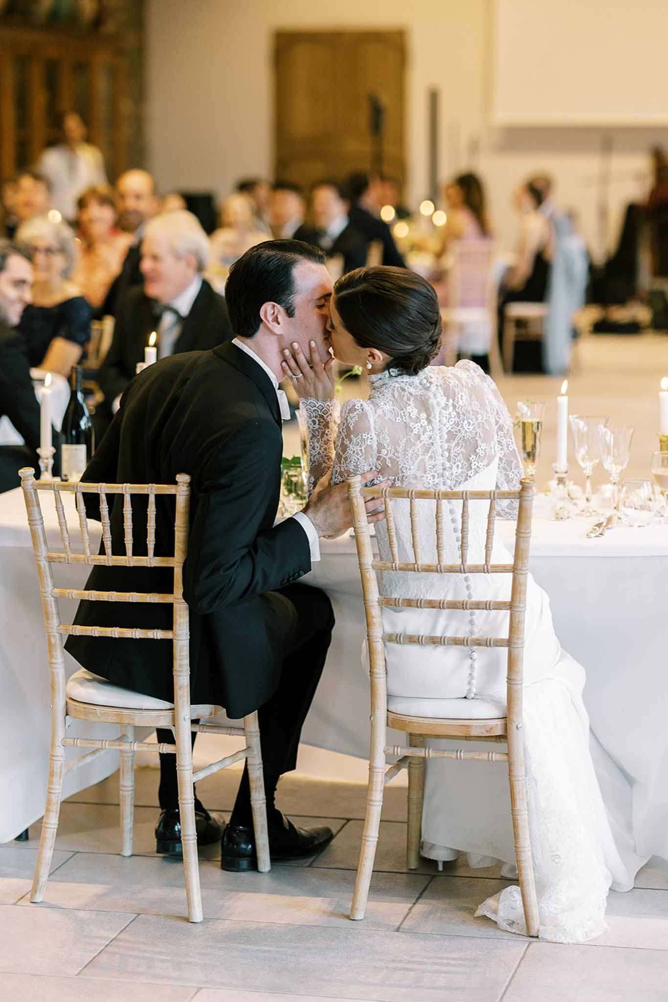 Bride and groom kissing at head table with taper candles and lace cape overlay during indoor reception