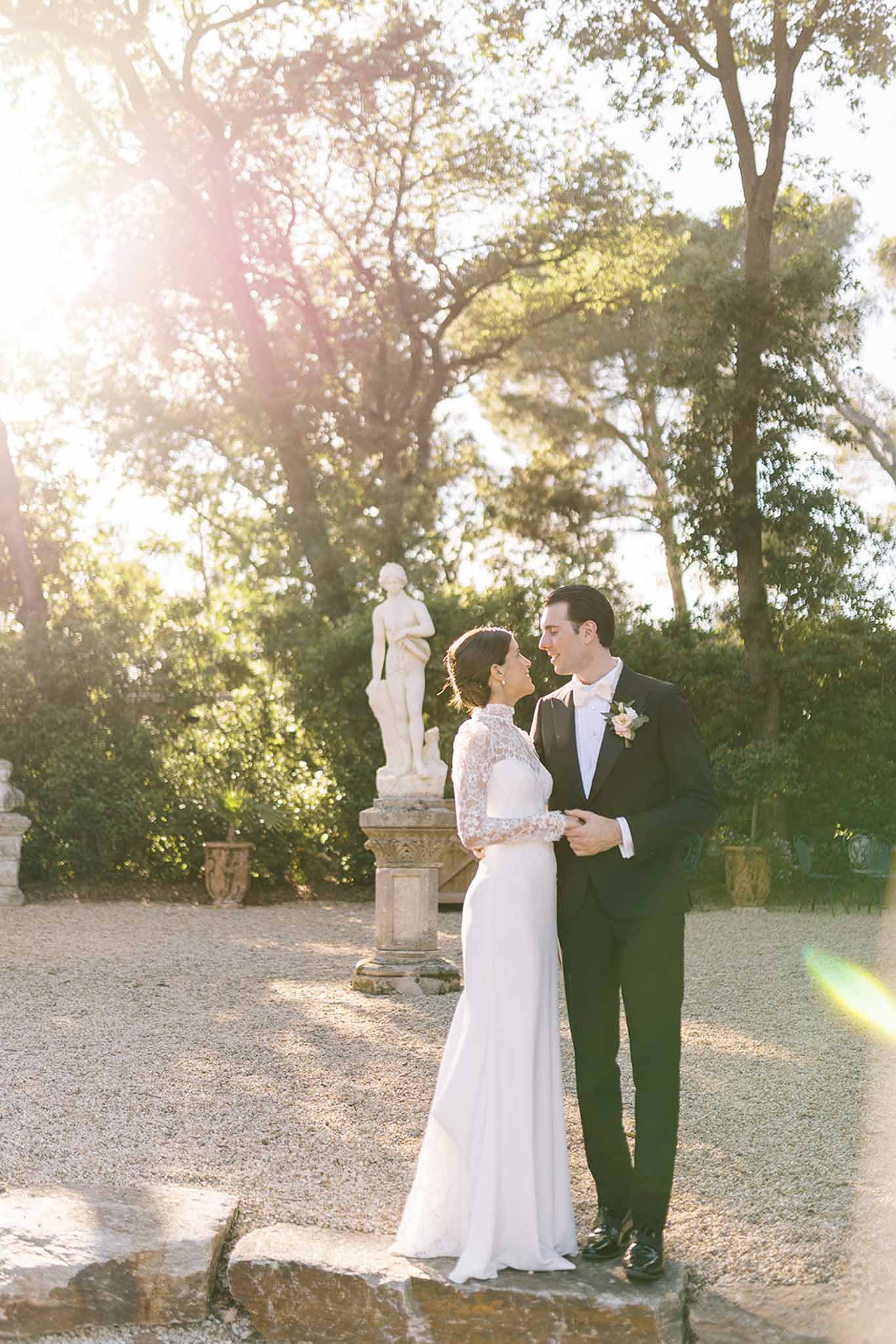 Couple smiling face-to-face at golden hour beside marble statue in formal chateau garden