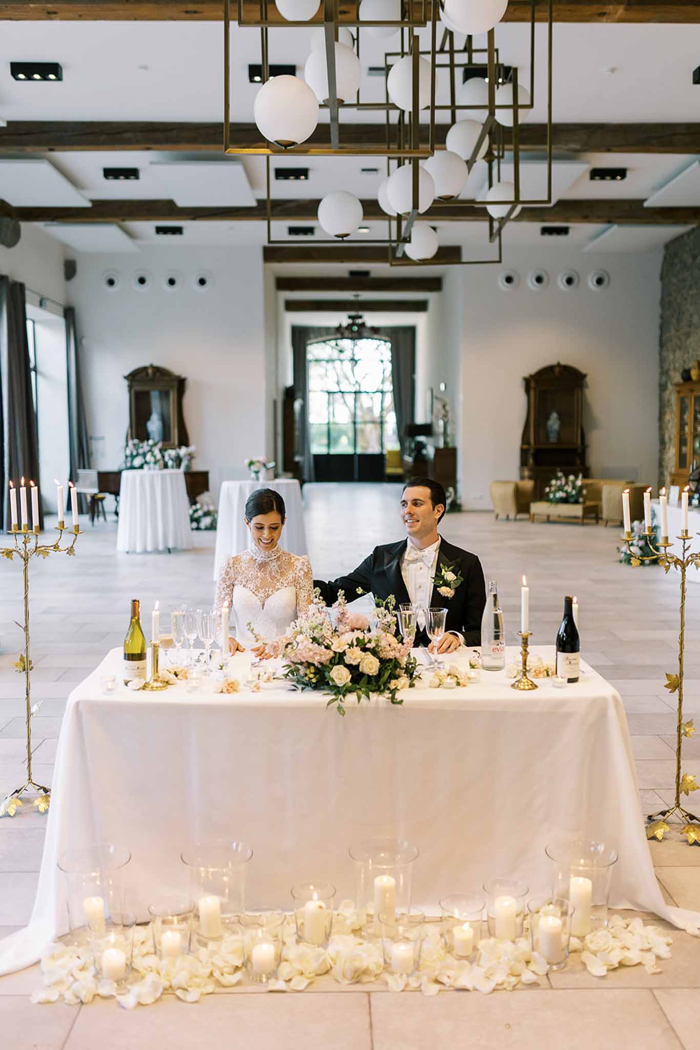 Couple at sweetheart table with blush roses, gold candelabra, and votive candles in beamed ballroom