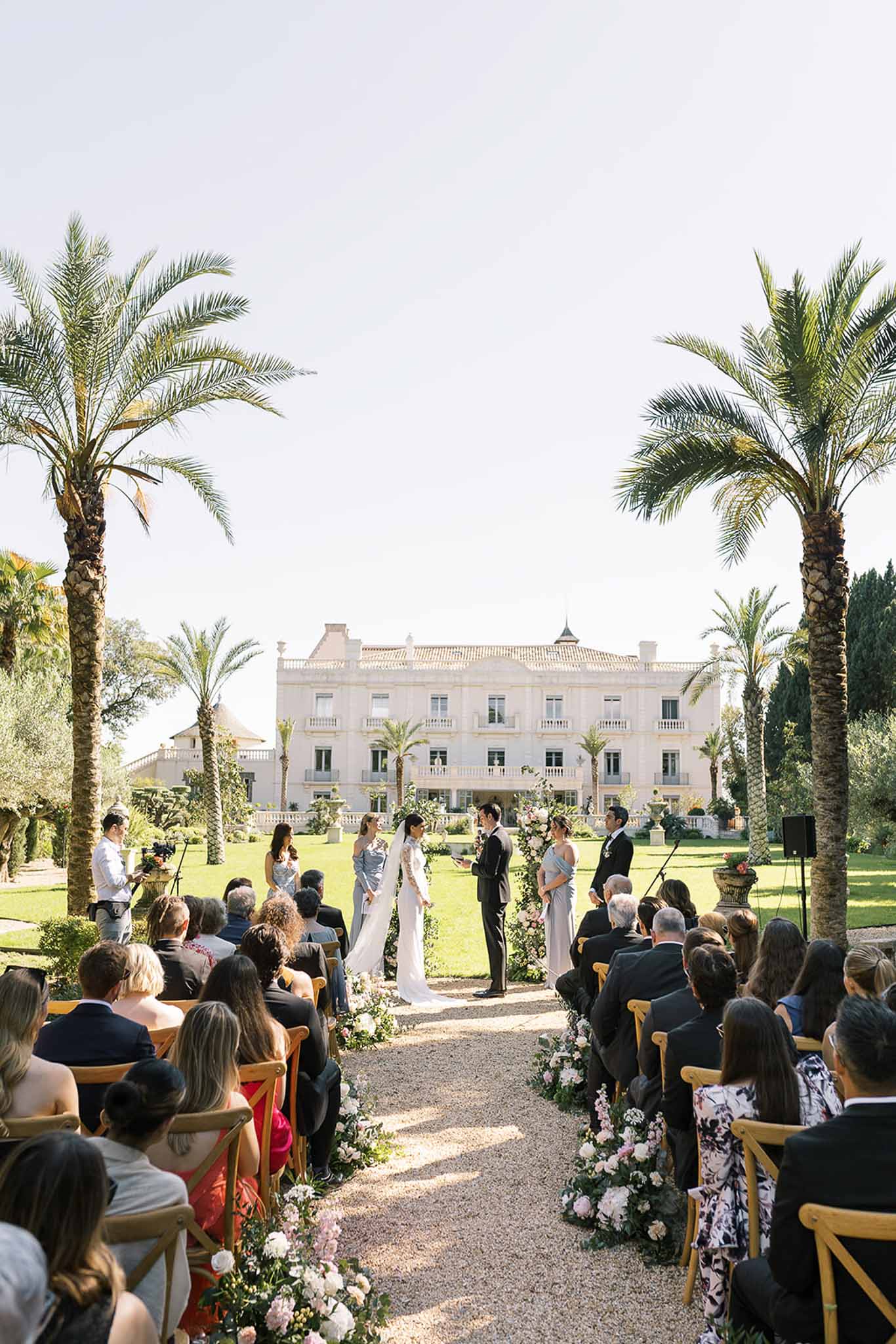 Sixty guests at garden ceremony before white Belle Epoque villa with cross-back chairs and palm trees framing aisle