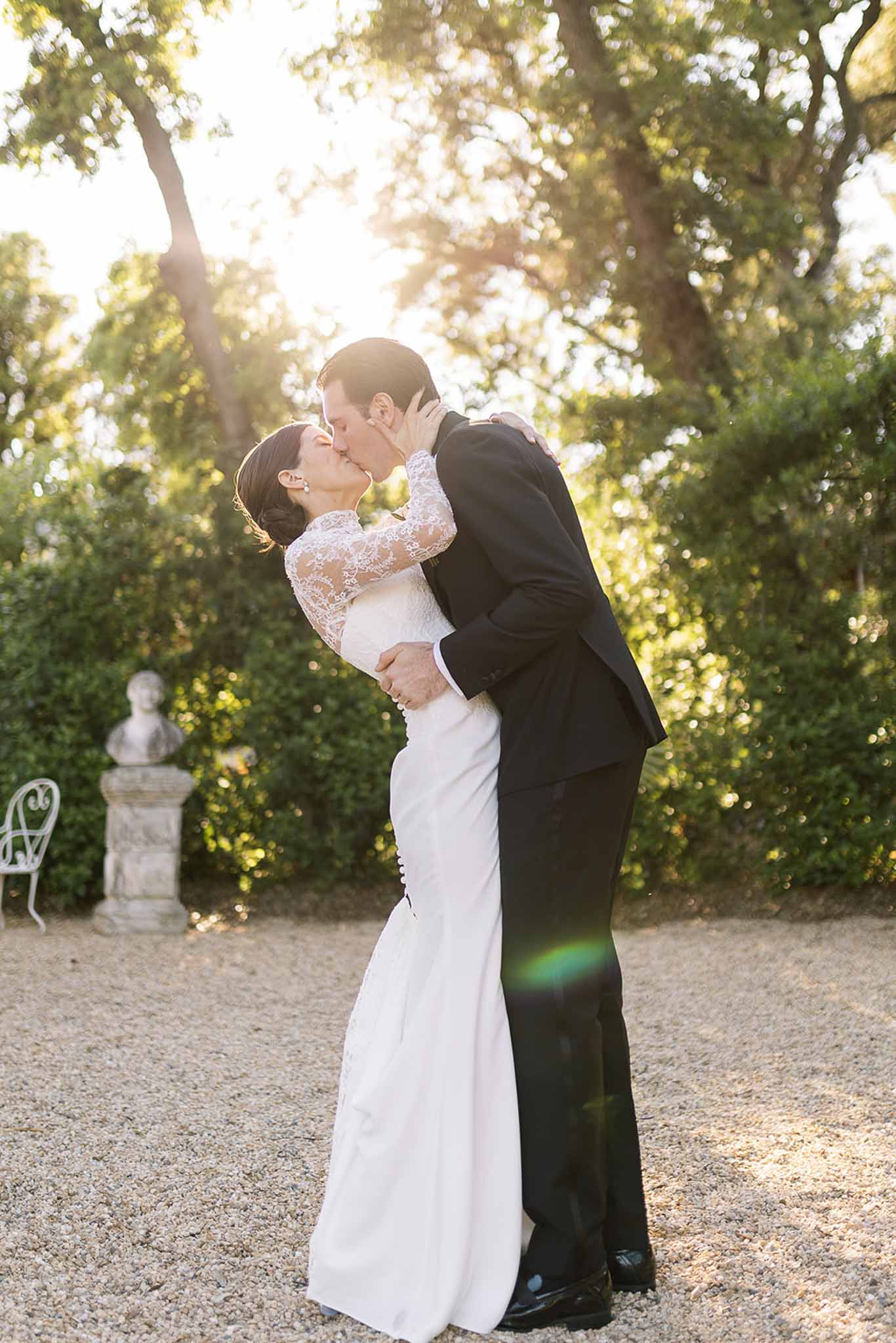 Dip kiss on gravel path with golden backlight through trees, bride in long lace-sleeve ivory gown