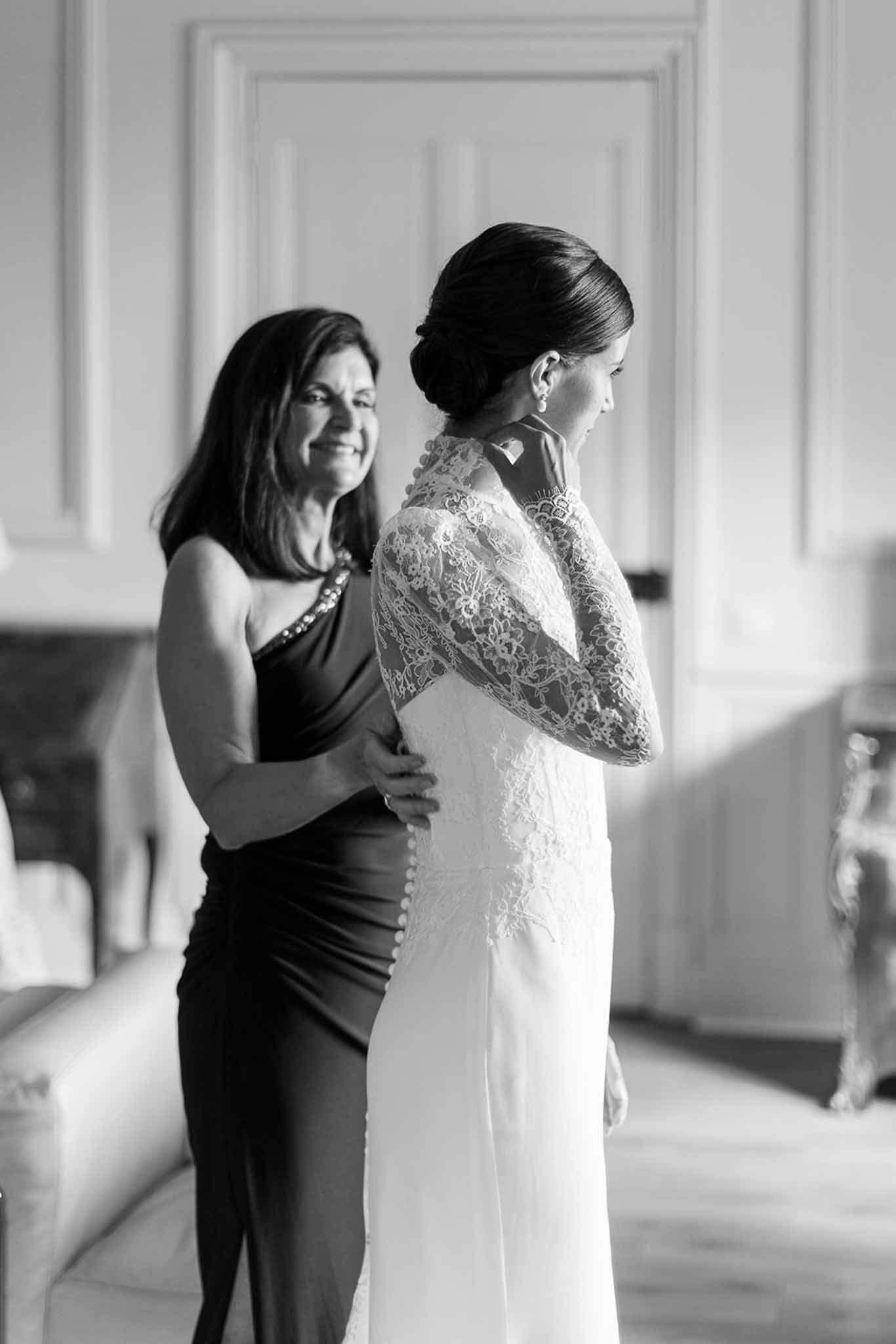 Mother fastening the back of the bride's long-sleeve lace gown in a wood-panelled room in black and white