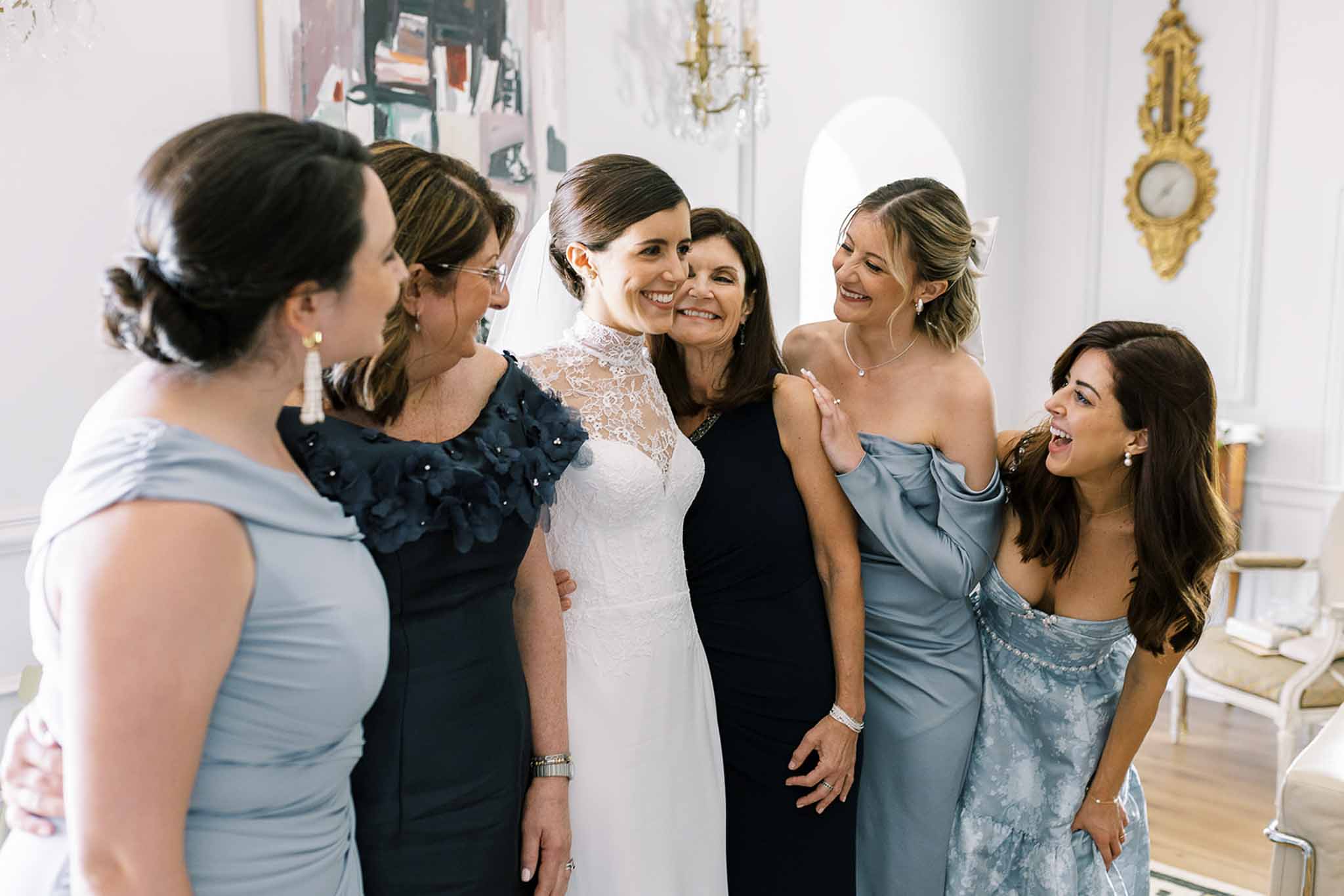 Bride in white lace gown with five women in coordinated blue and navy dresses in chandelier room