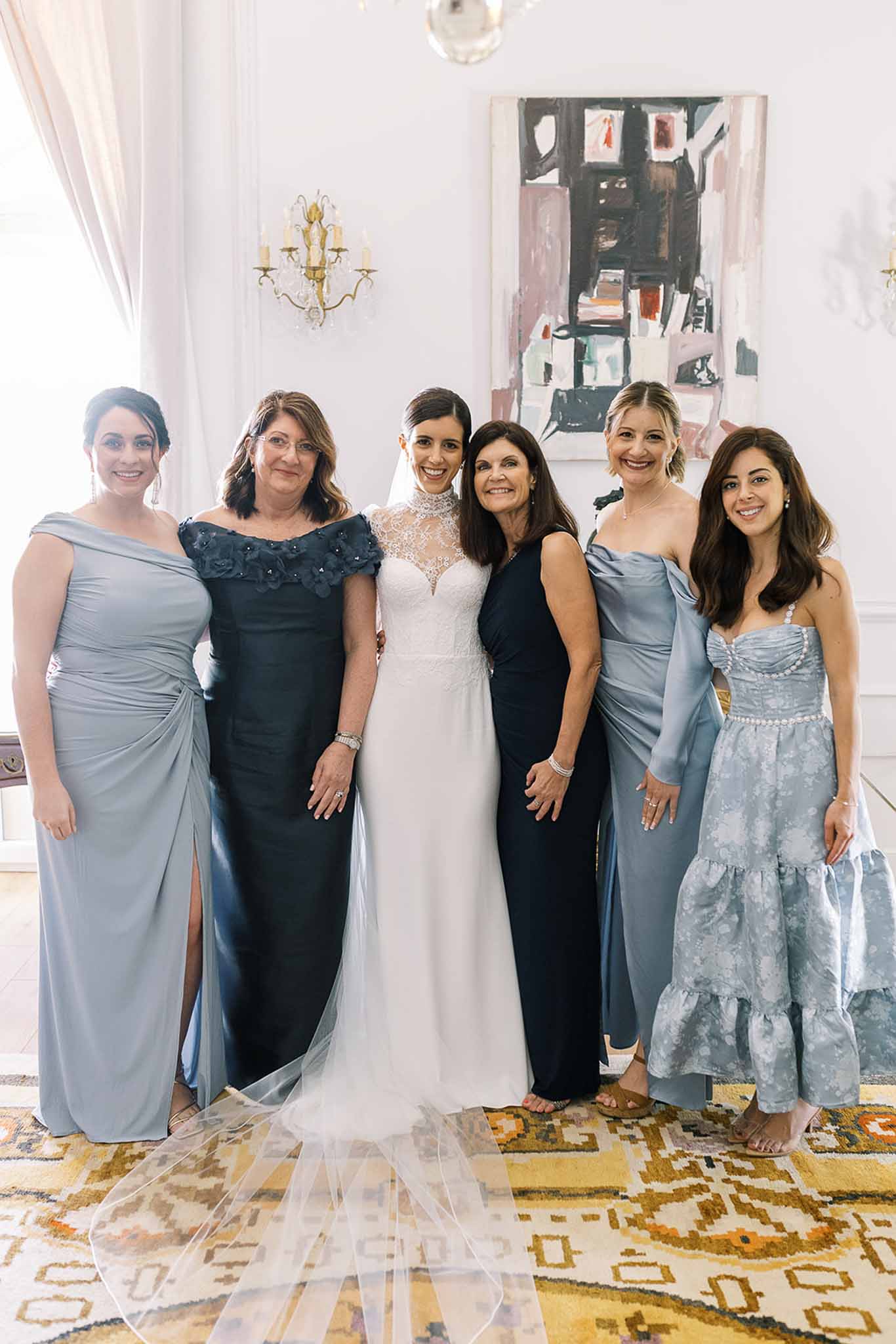Bride in white lace gown with five women in dusty blue to navy dresses in bright interior with abstract painting