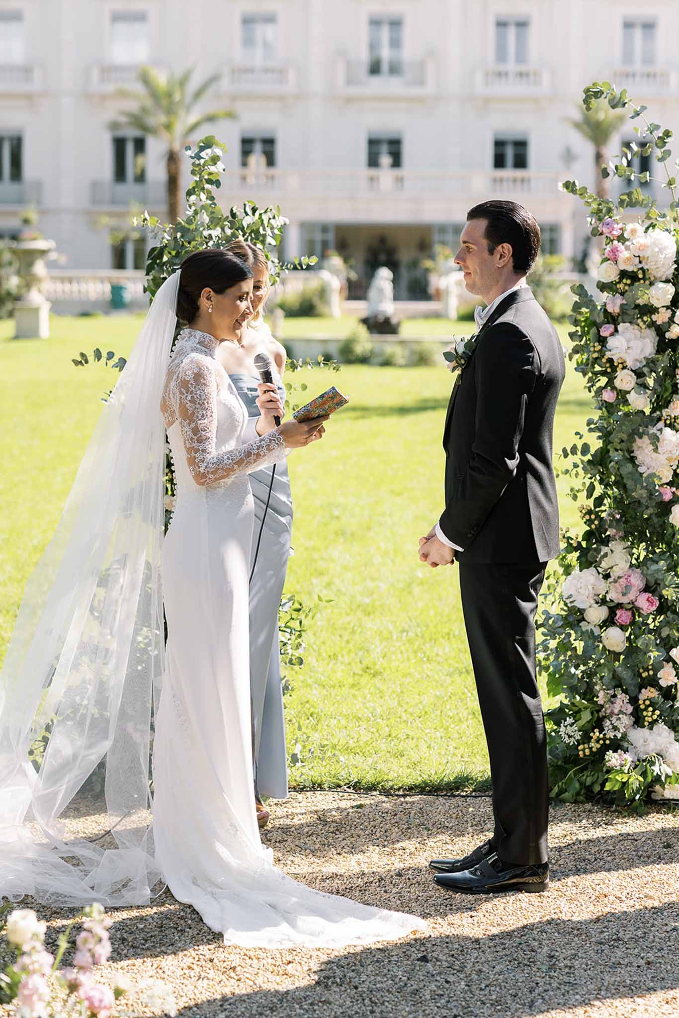 Bride reading vows to groom at outdoor chateau ceremony with rose and hydrangea floral arch
