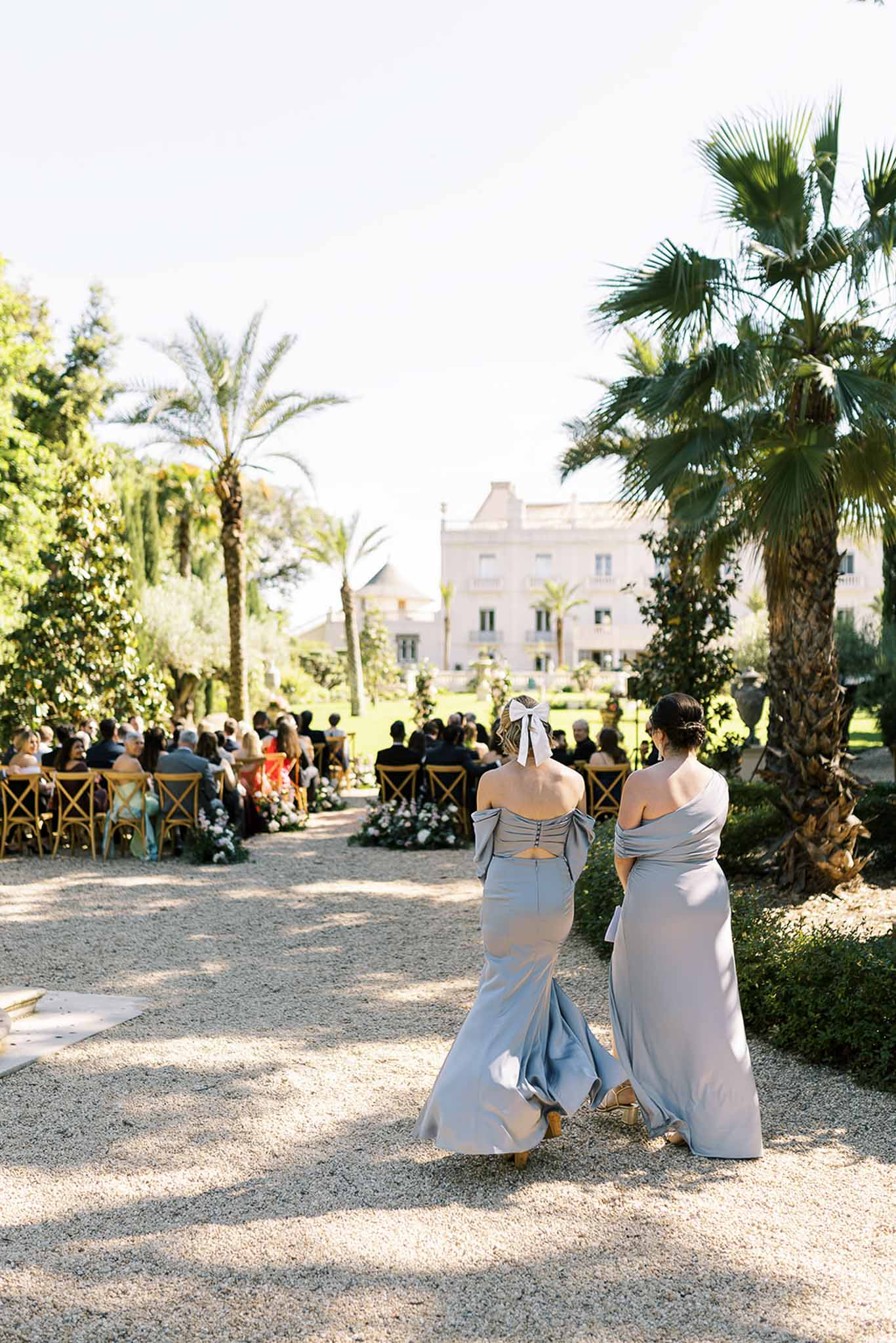 Two bridesmaids in powder blue satin gowns walking toward outdoor ceremony with seated guests and floral arch