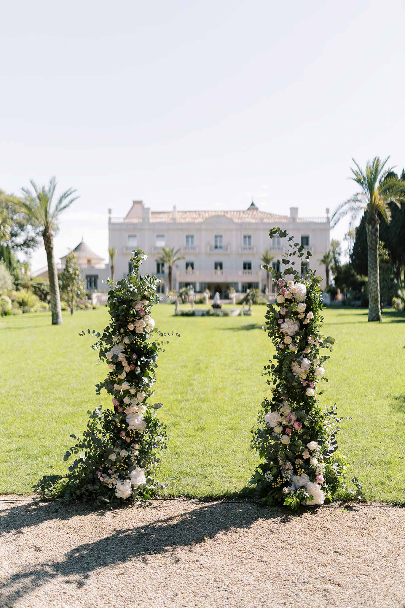 Two tall floral columns with roses and eucalyptus flanking gravel path to neoclassical chateau