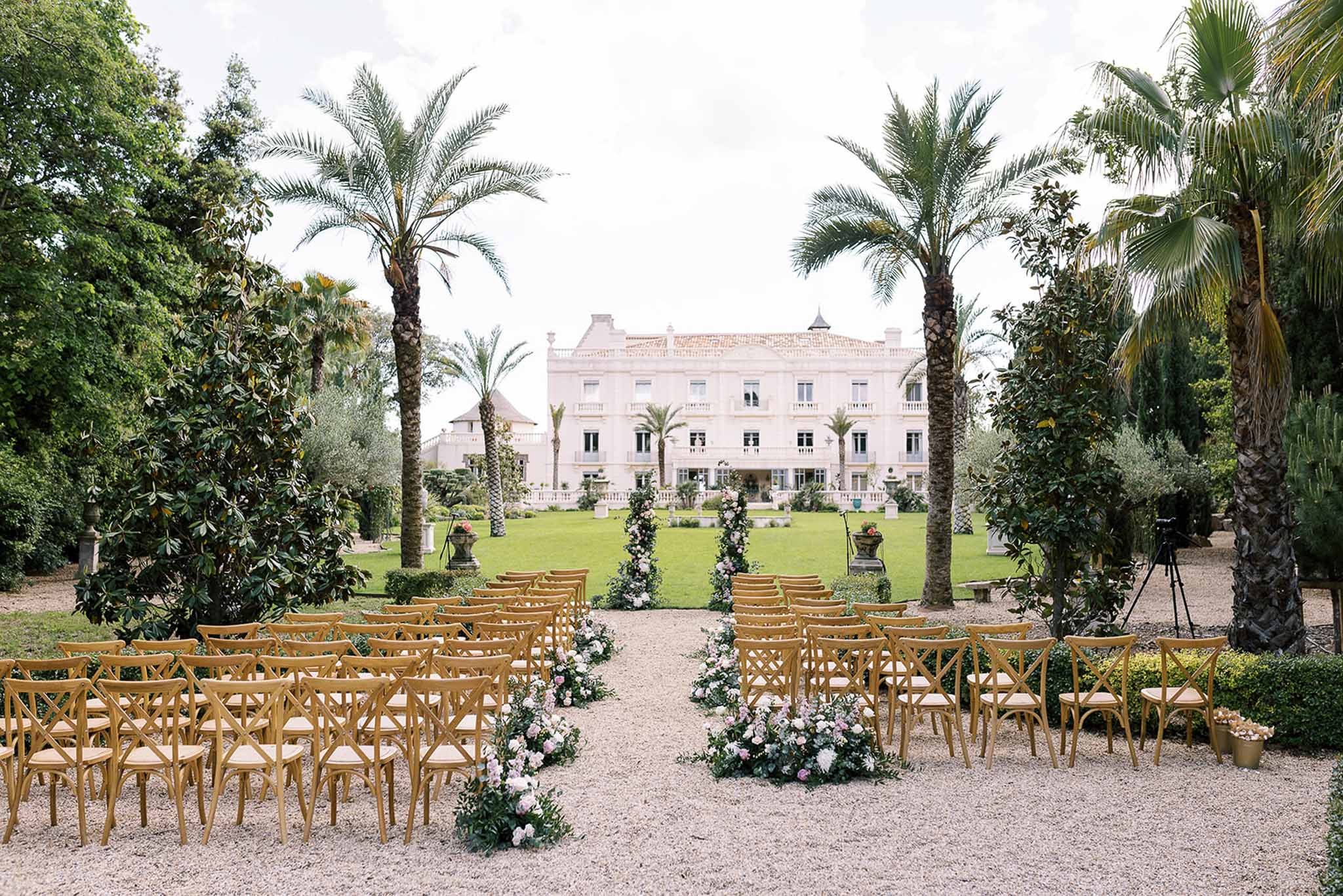 Outdoor ceremony setup with wooden cross-back chairs and blush floral aisle arrangements on gravel terrace before white manor