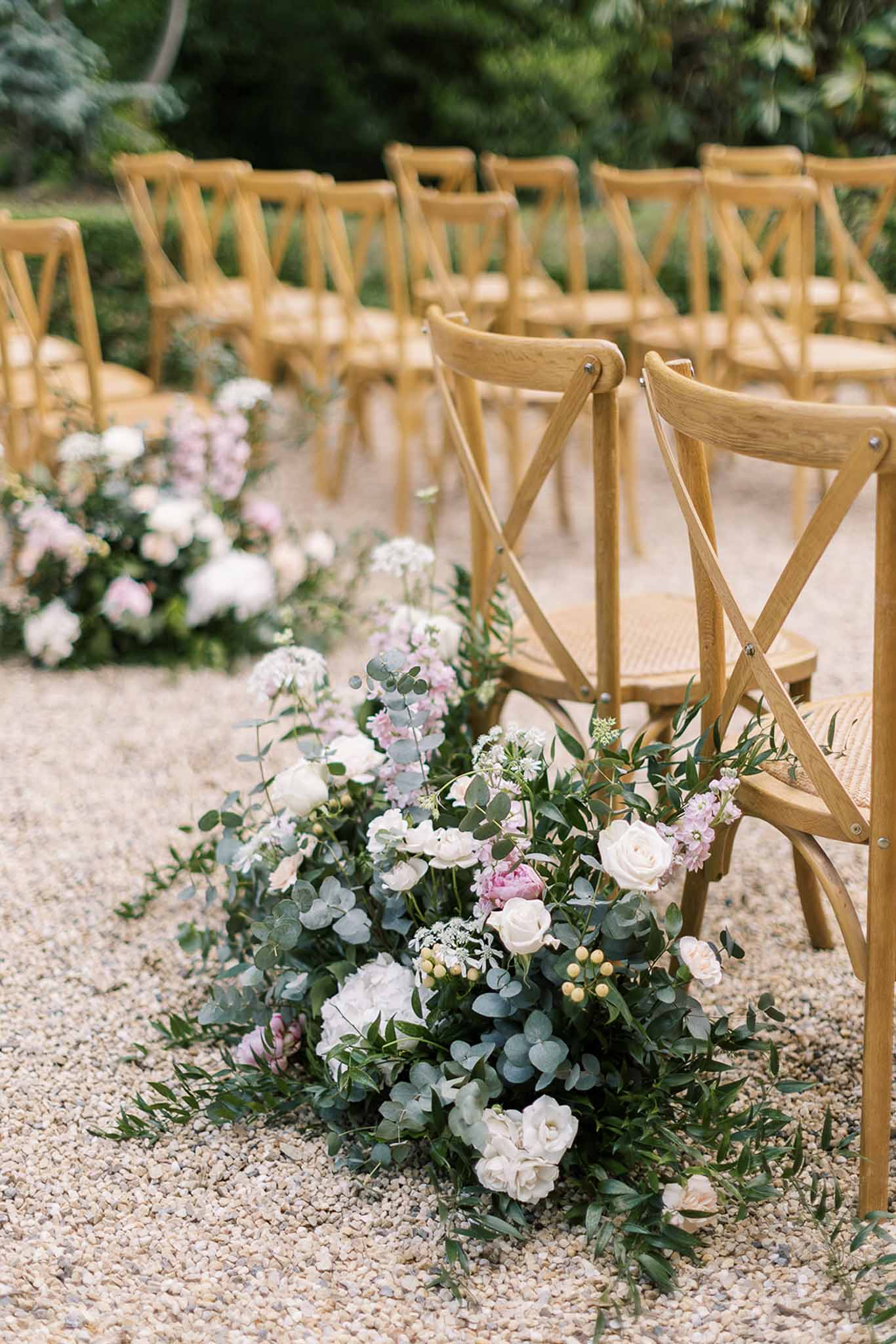Aisle ground clusters of ivory roses, blush stock, and eucalyptus beside oak cross-back chairs on gravel