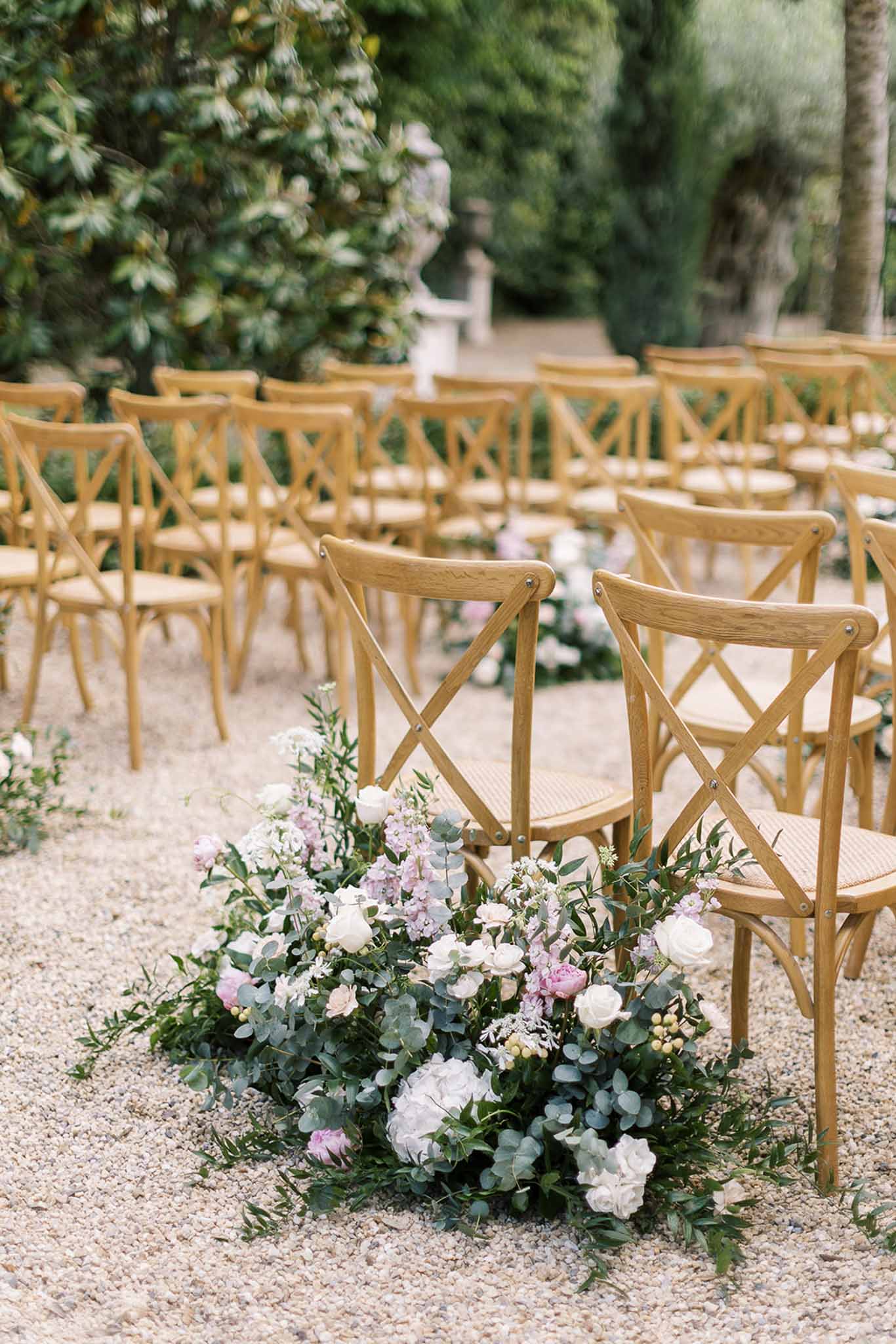 Low-angle blush peony, white rose, and mauve stock aisle arrangement with cross-back chairs on gravel