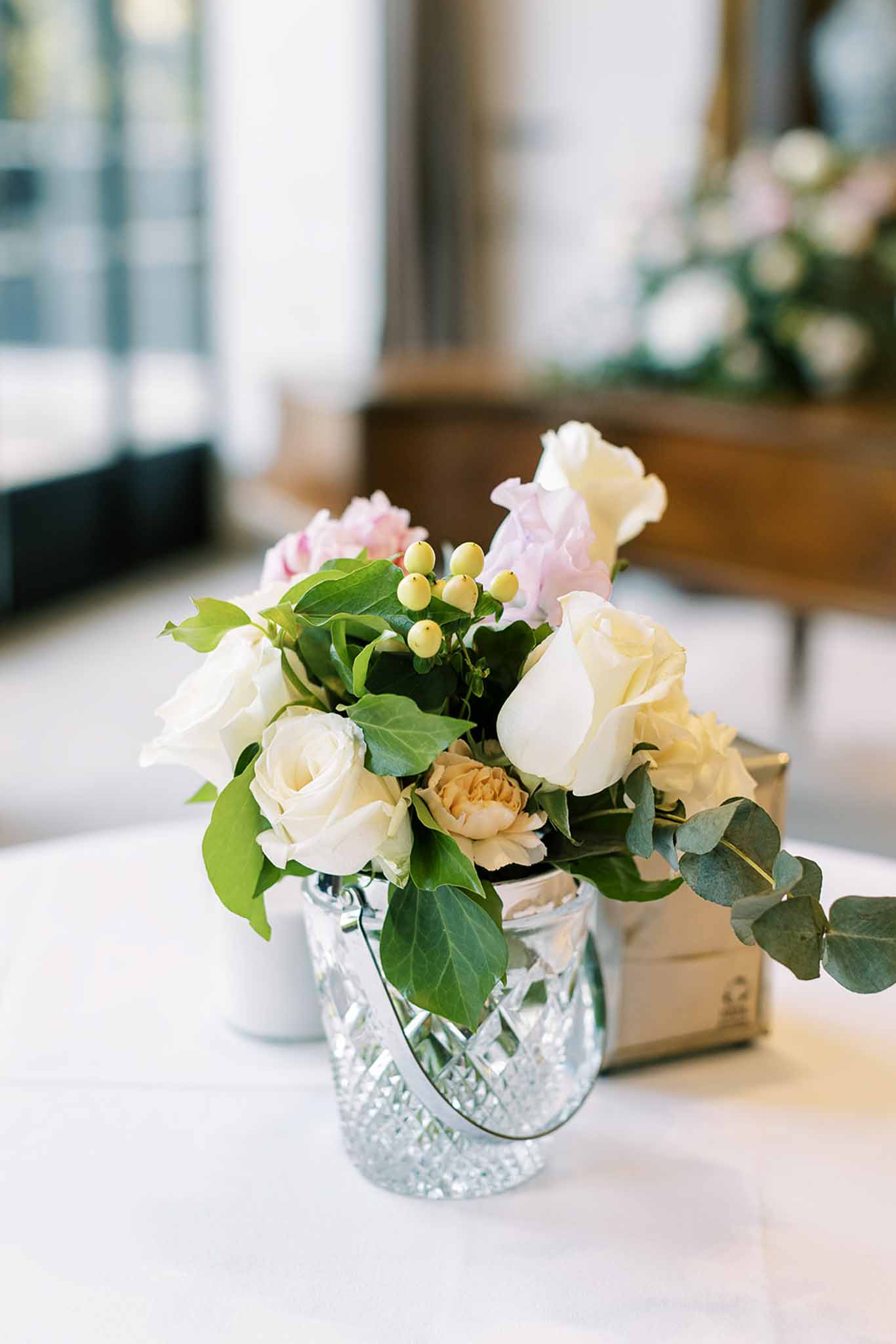 Crystal bucket vase with blush garden roses, peach ranunculus, and eucalyptus on white linen