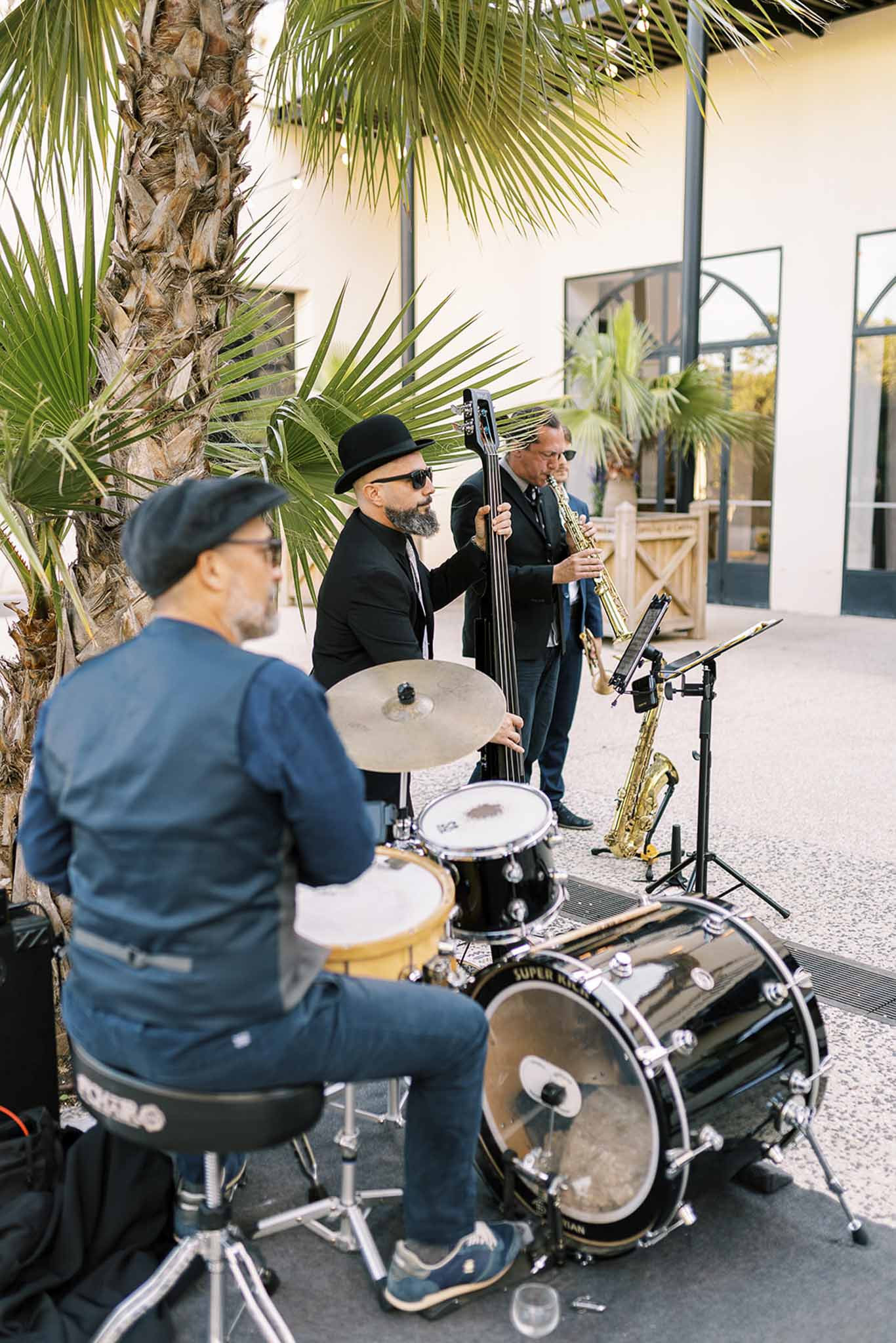 Live jazz trio with drums, upright bass, and saxophone performing on venue terrace beside palm tree