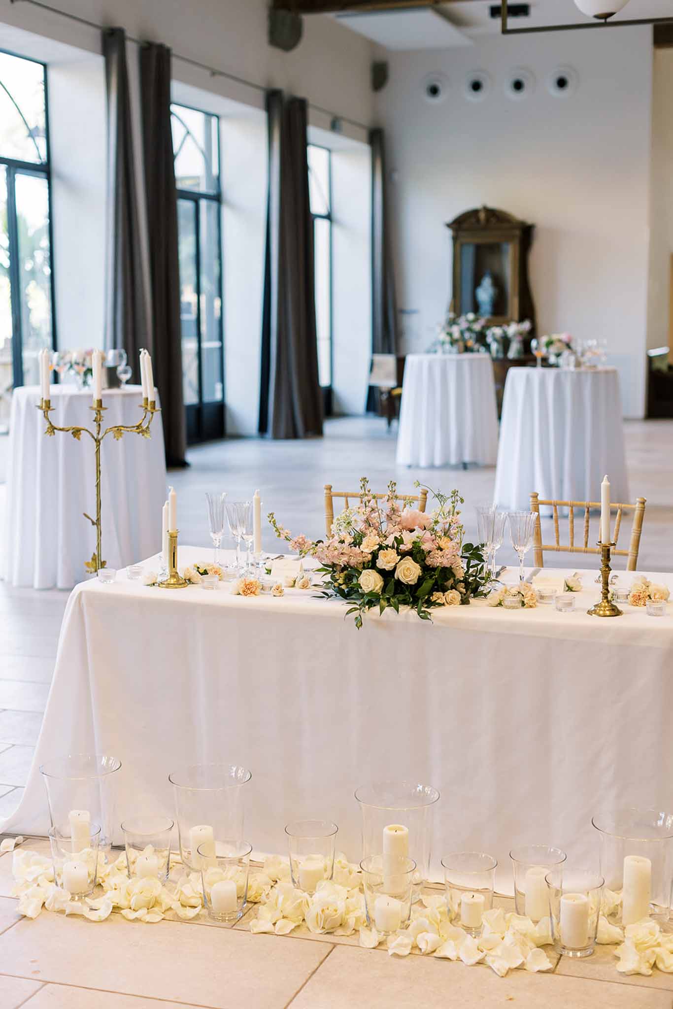 Indoor sweetheart table with blush and cream roses, gold chiavari chairs, and taper candles in a bright ballroom