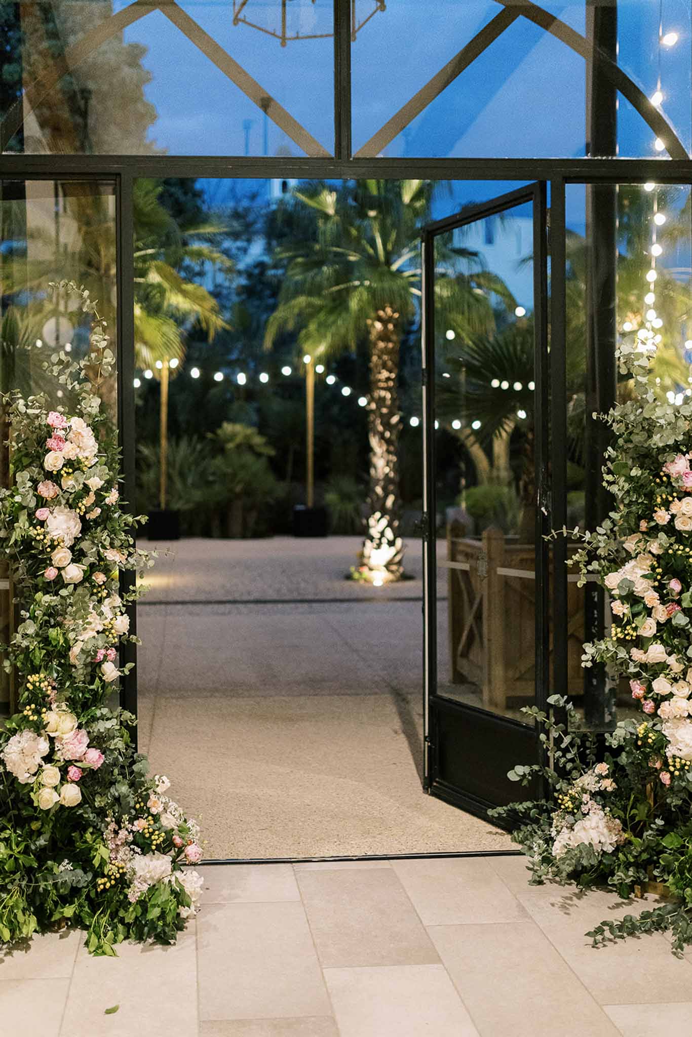 Glass-door entrance with blush rose floral columns and palm-tree festoon courtyard at blue hour