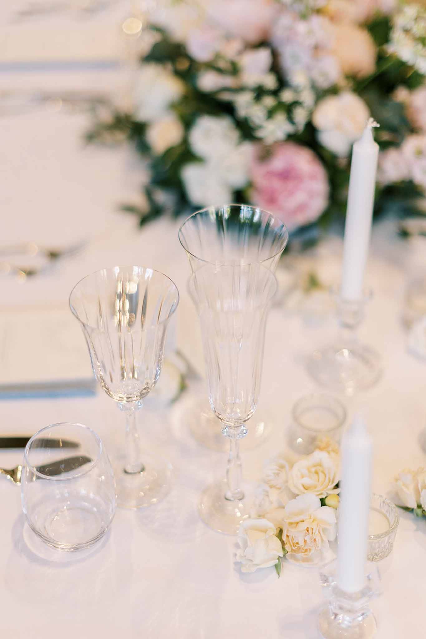 Crystal wine glasses and taper candles beside blush garden roses on white linen reception table