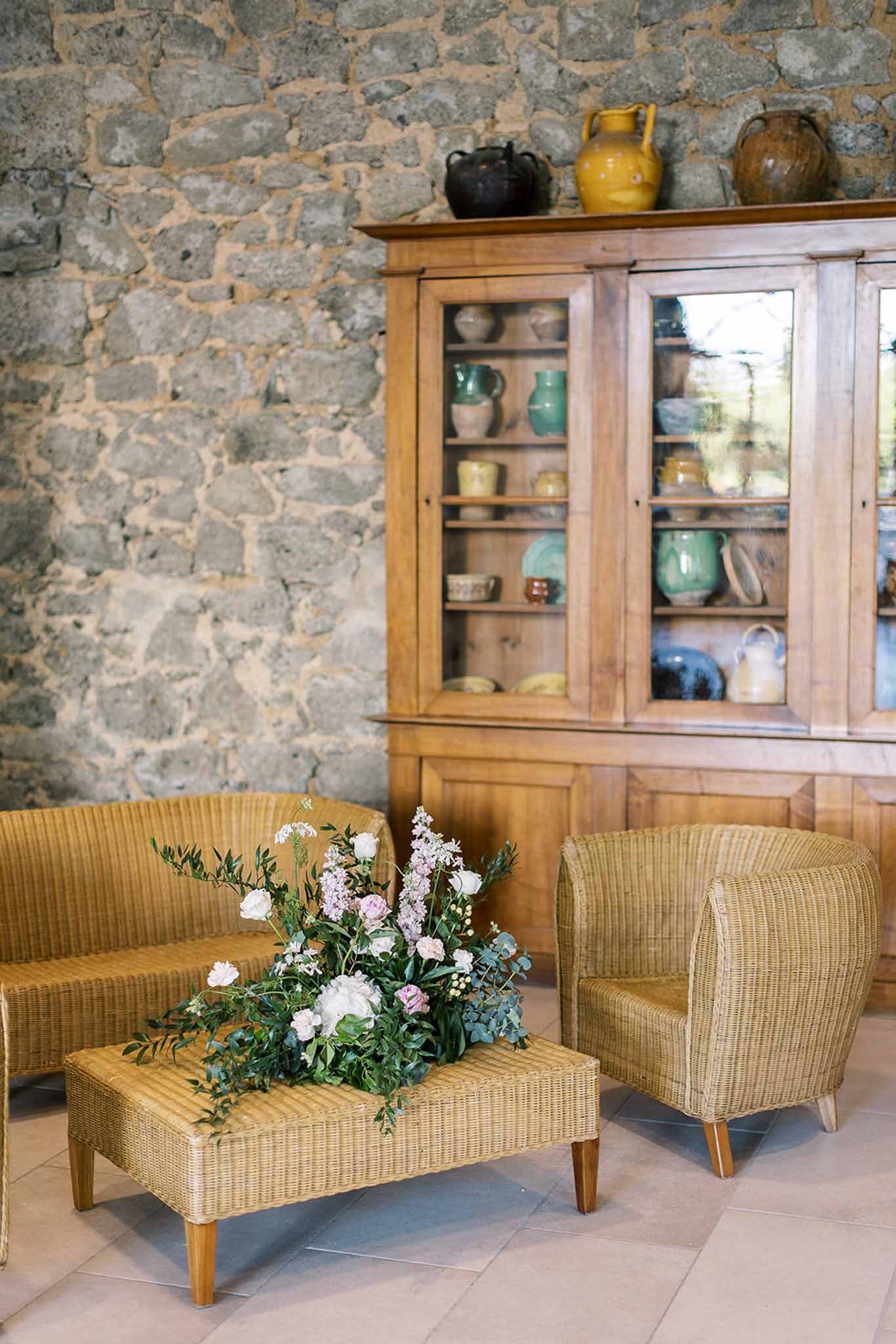 Rustic reception lounge with wicker furniture, white peonies and blush floral arrangement, exposed stone walls