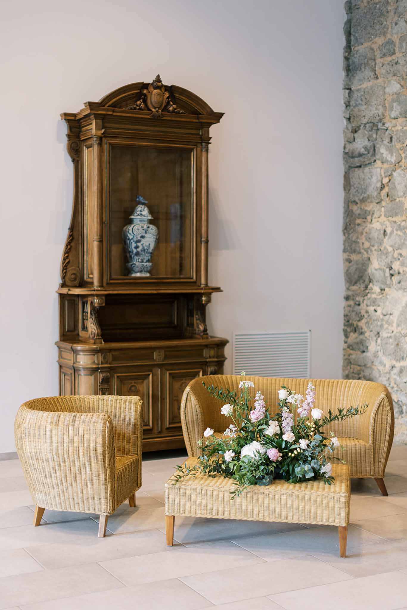Wicker loveseat with white rose and blush delphinium arrangement beside carved walnut cabinet