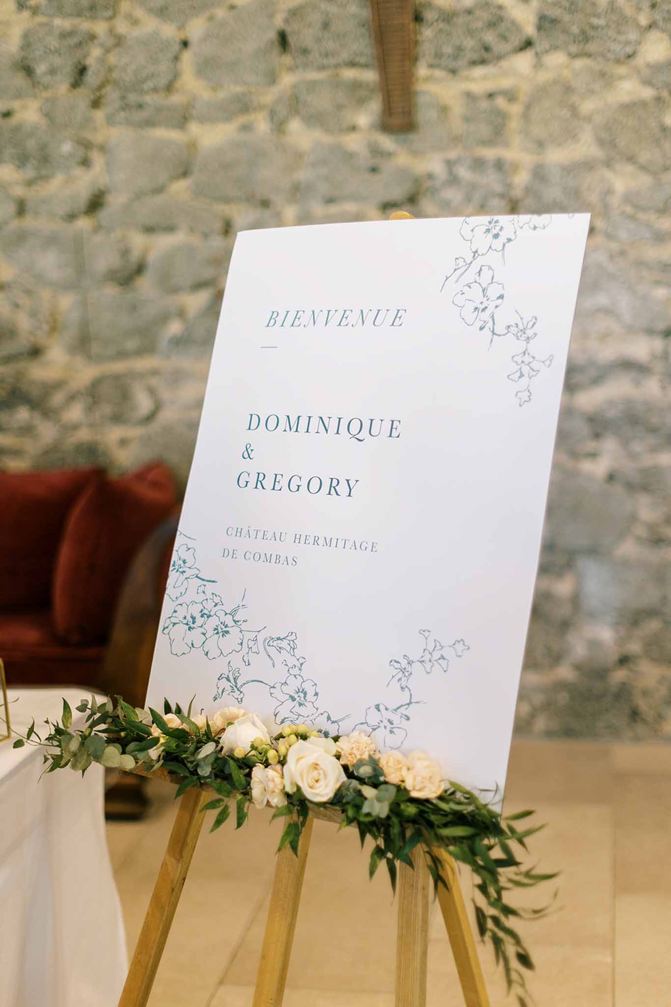 Bienvenue welcome sign on gold easel with teal botanical illustrations and cream rose arrangement at base