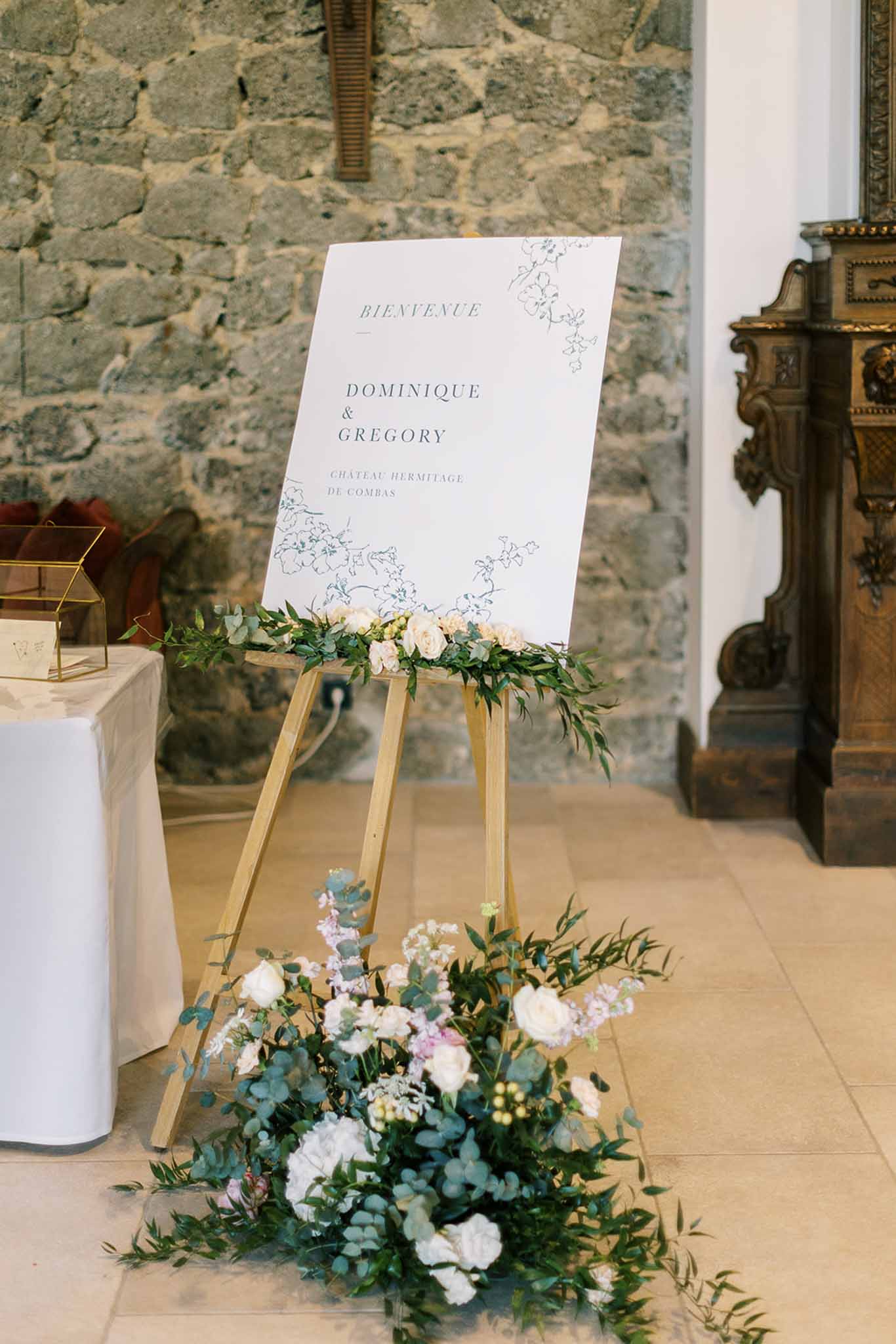 White welcome sign on wood easel with blush rose garland and white floral floor arrangement in chateau stone interior