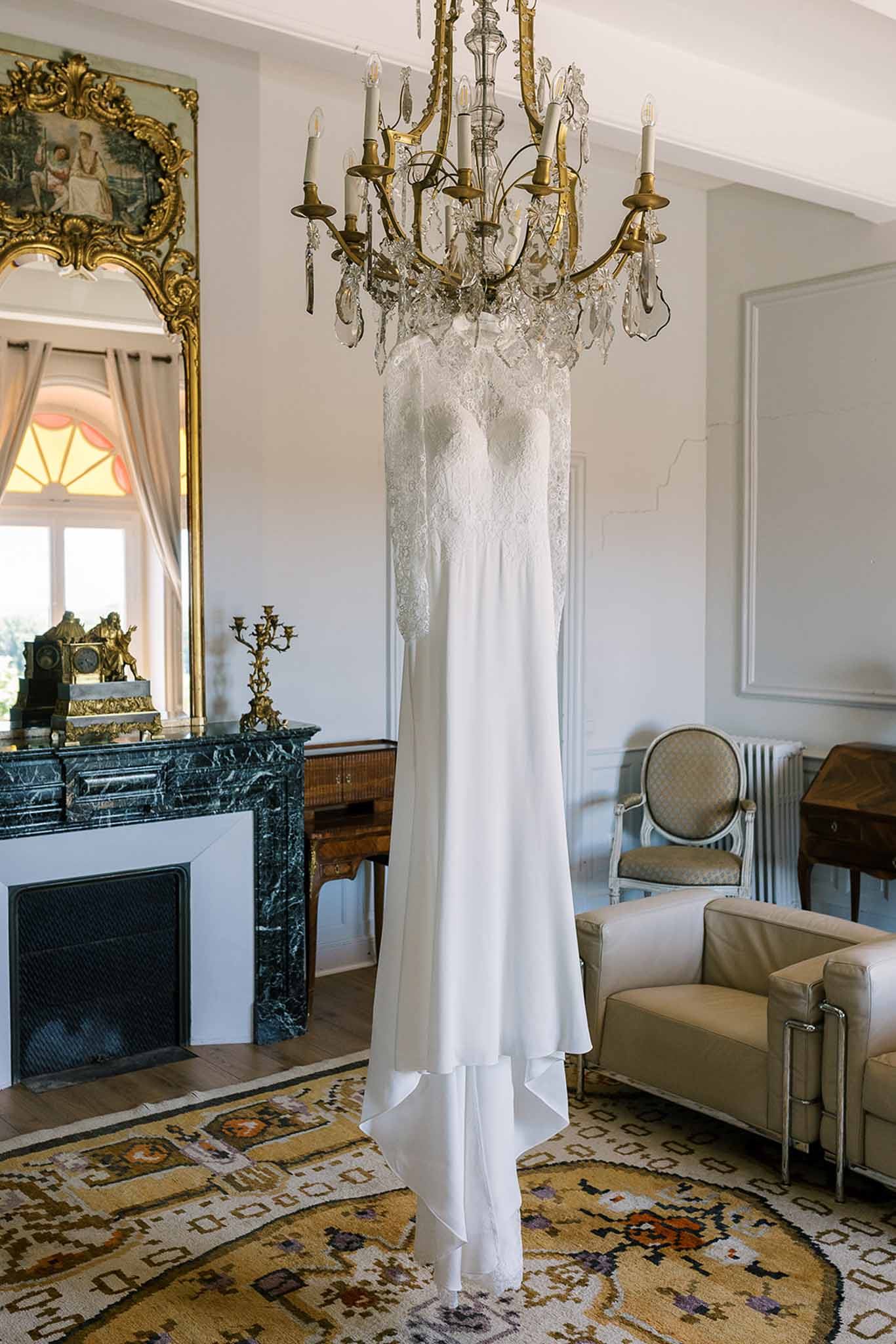Lace-bodice crepe gown hanging from chandelier in chateau room with green marble fireplace and gilt mirror
