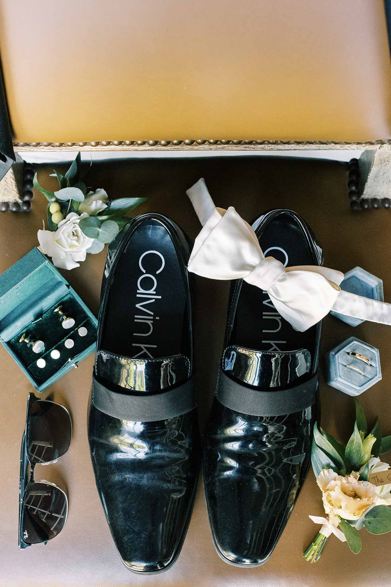 Groom flat lay with black patent loafers, ivory bow tie, teal ring boxes, gold cufflinks, and boutonniere