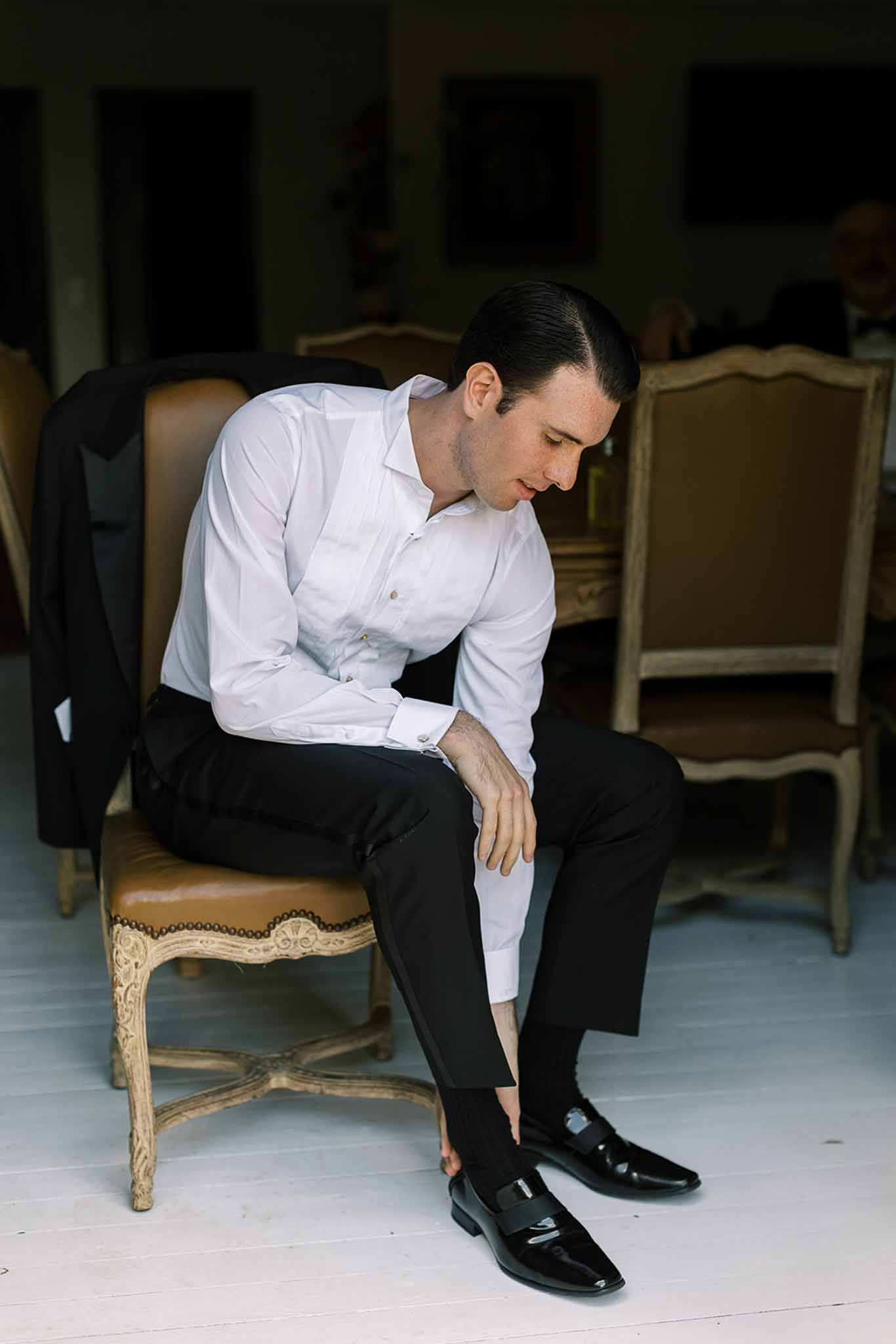 Groom seated adjusting black patent shoes with tuxedo shirt and jacket draped on French chair