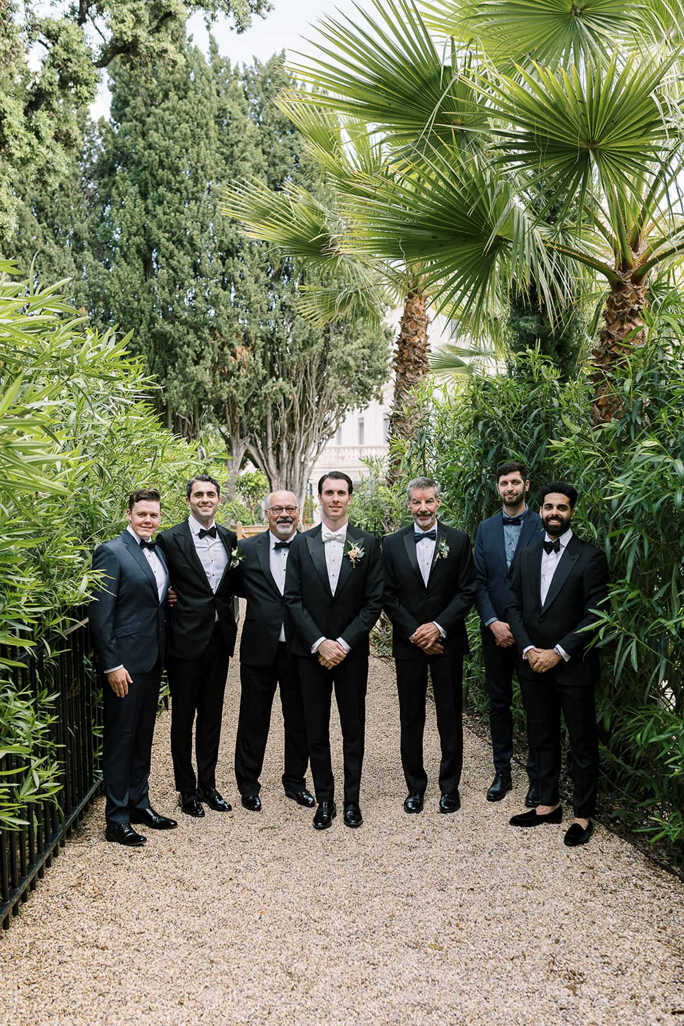 Groom and six groomsmen in black tuxedos with white bow ties on gravel path with palm trees