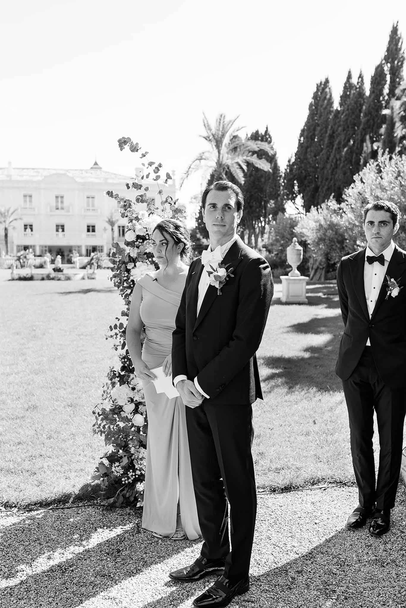 Groom at altar with attendant and asymmetric floral installation before pale estate in B&W