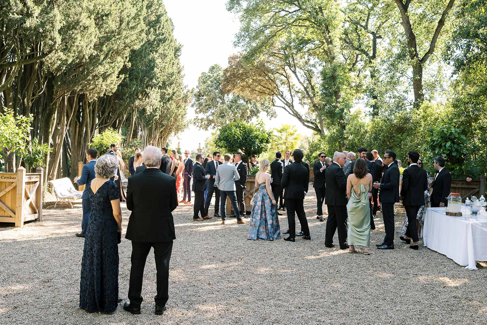 Thirty guests in formal attire mingling on gravel terrace at French estate garden during afternoon cocktail hour