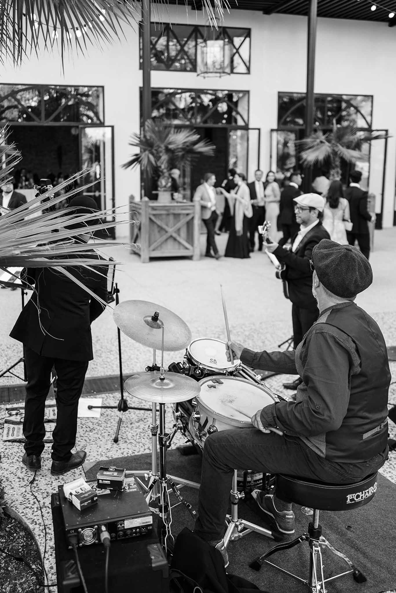 Black and white view from behind drummer performing for formal guests at industrial venue with palm trees and lanterns
