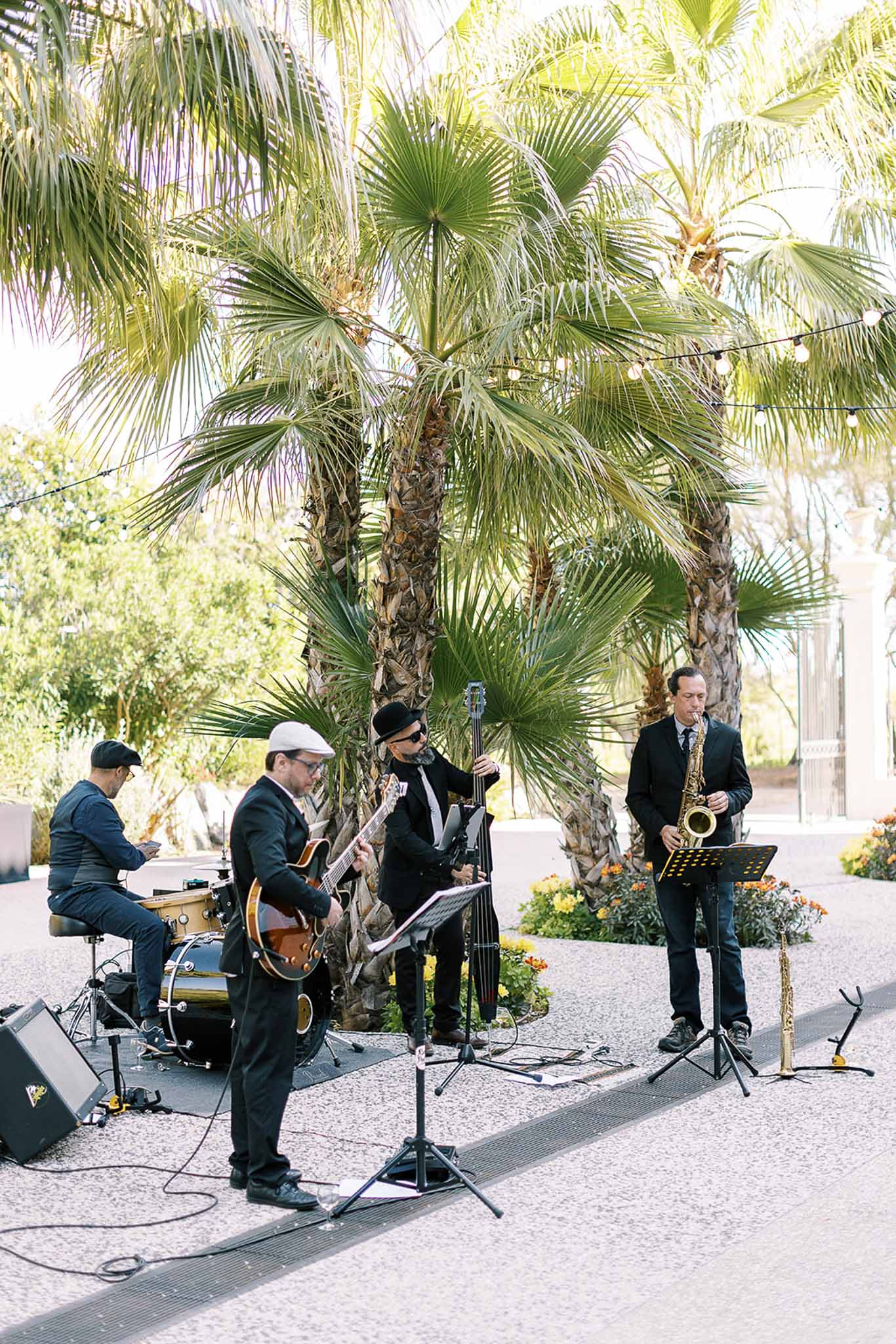 Four-piece jazz band in black attire performing on gravel terrace with festoon lights overhead