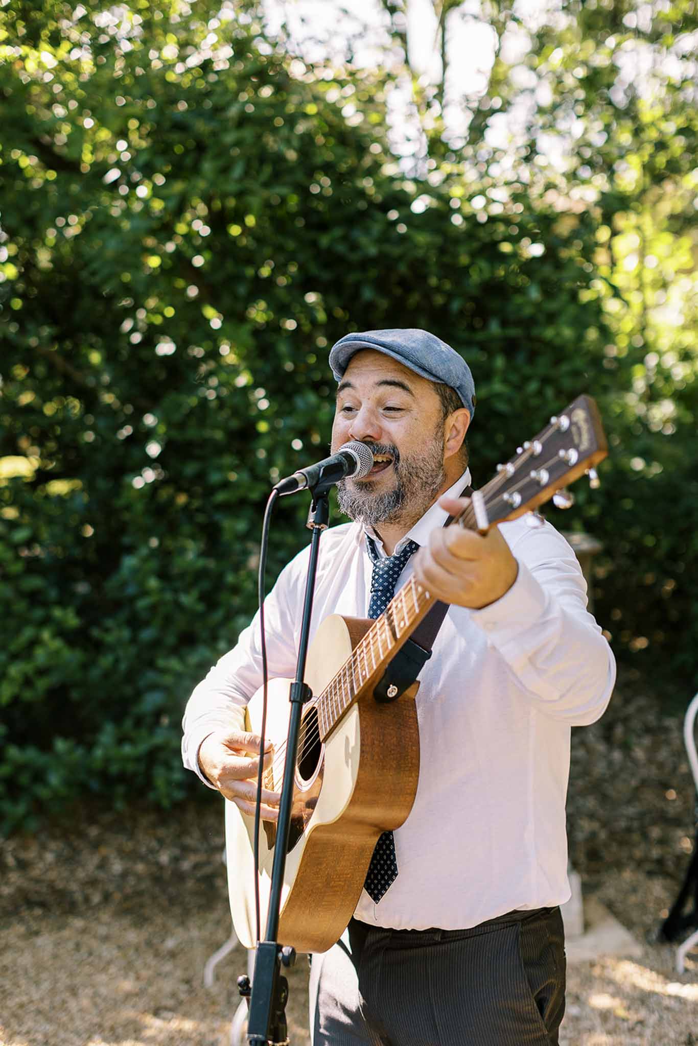 Musician in flat cap playing acoustic guitar and singing at outdoor wedding reception