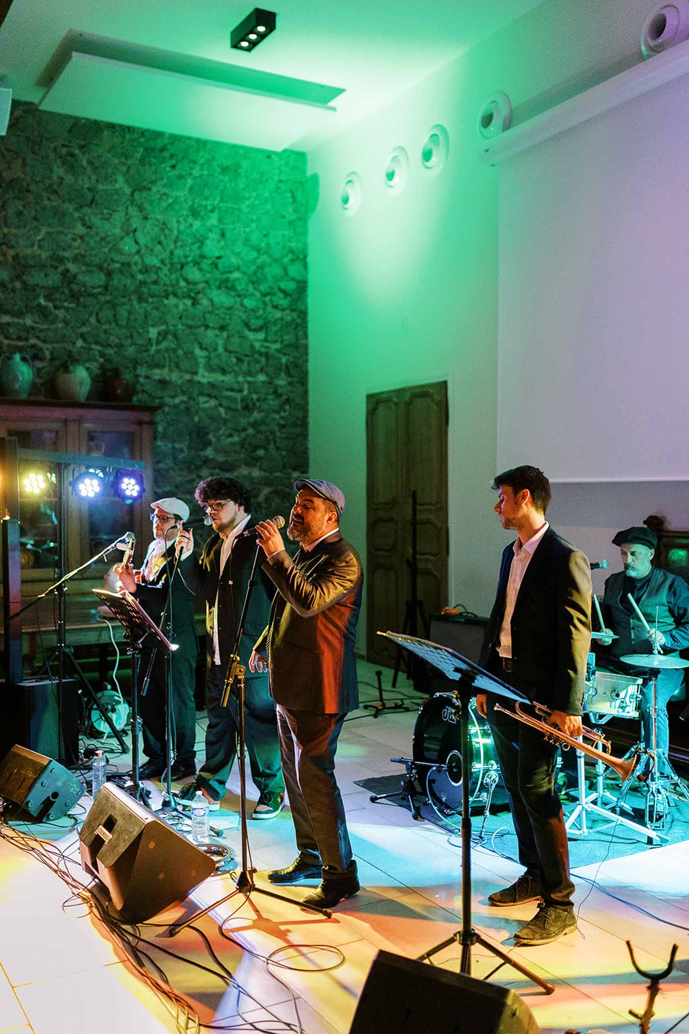 Five-piece wedding band performing in stone-walled venue with green uplighting, trumpet player and drummer visible on stage