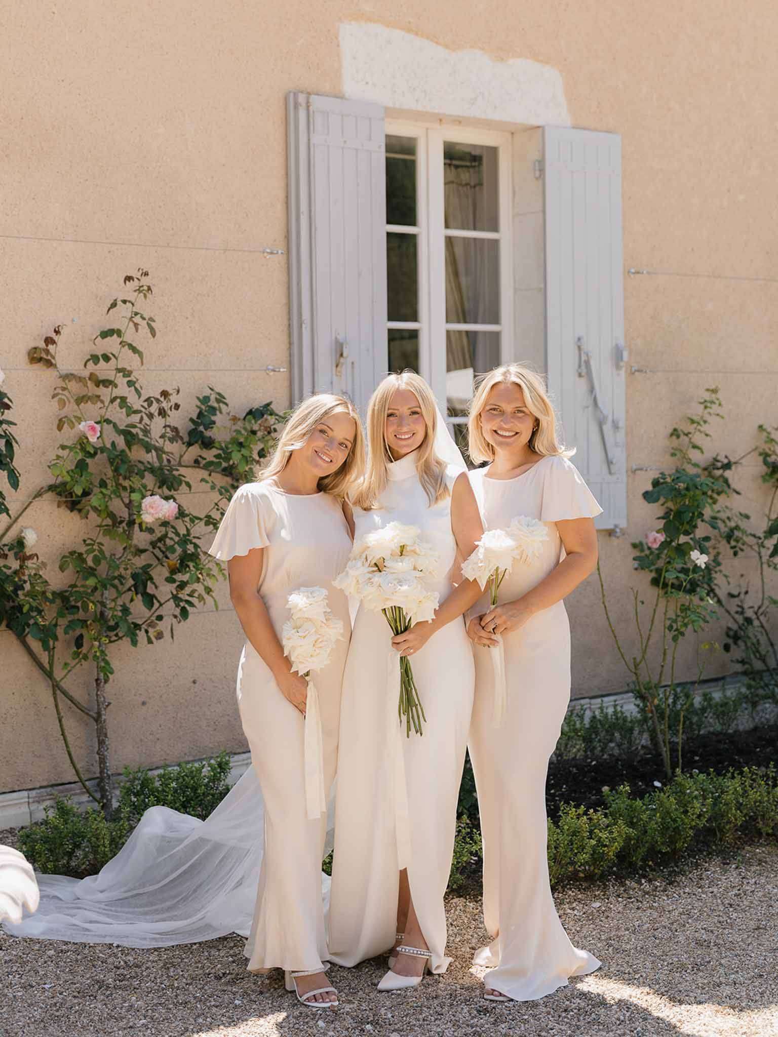 Bride and two bridesmaids in champagne satin dresses holding ivory peony bouquets before ochre manor
