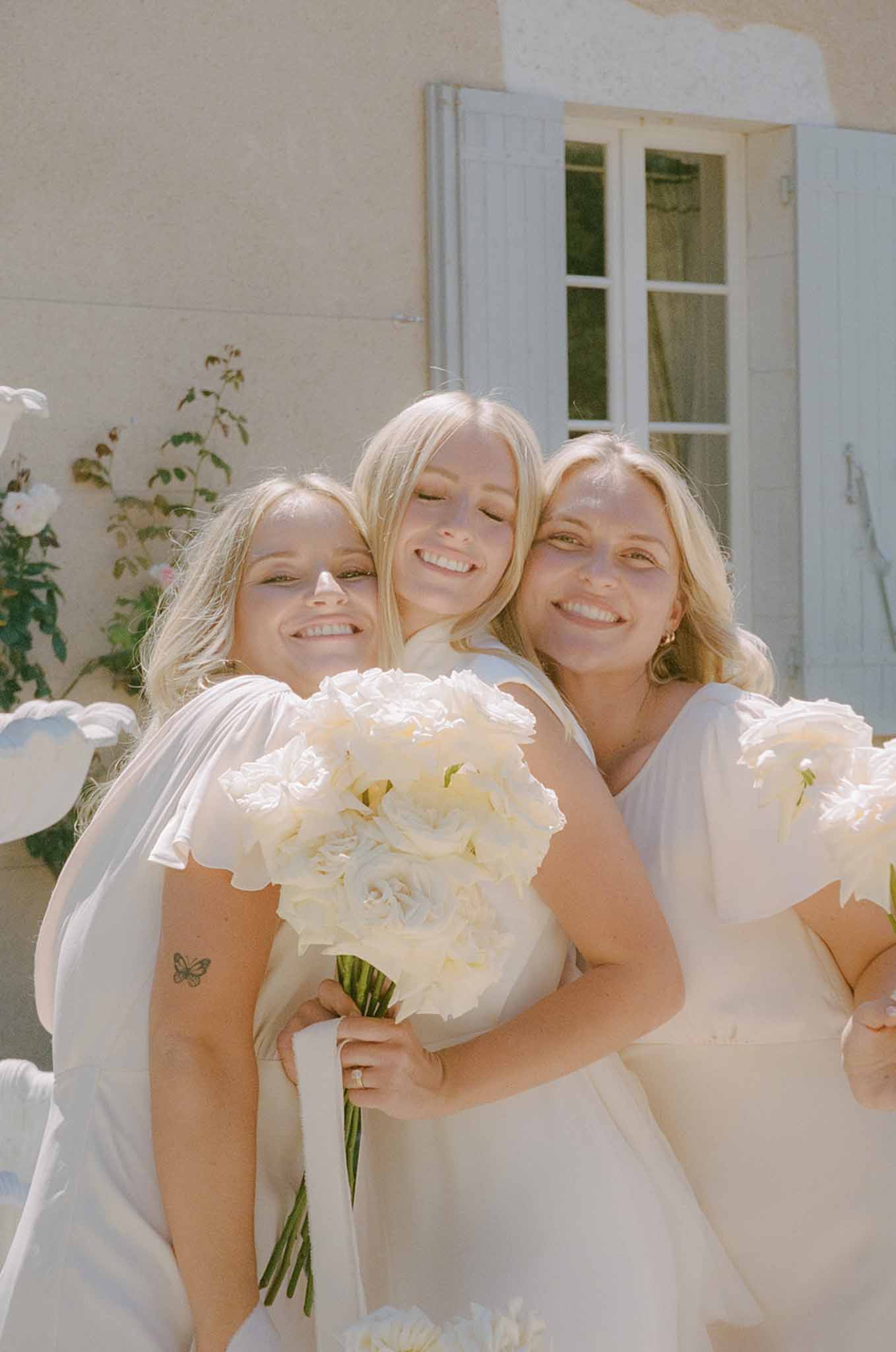 Bride and two bridesmaids in white dresses smiling together holding ivory peony bouquets outside stone building