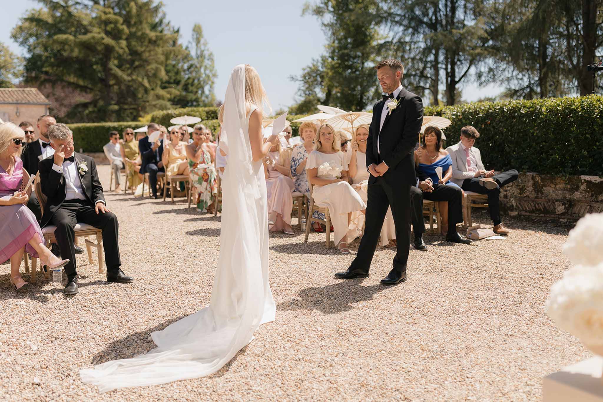 Bride reading vows to groom on gravel courtyard with guests holding cream parasols on gold chairs