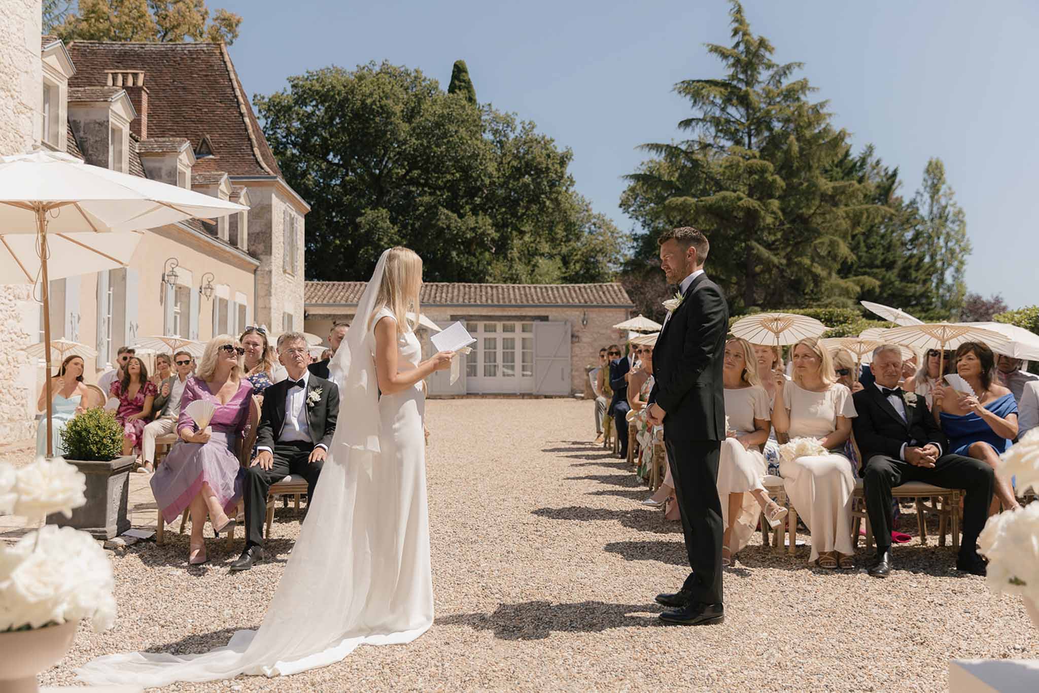 Outdoor ceremony on chateau courtyard with bride reading vows, guests with parasols, and white hydrangeas
