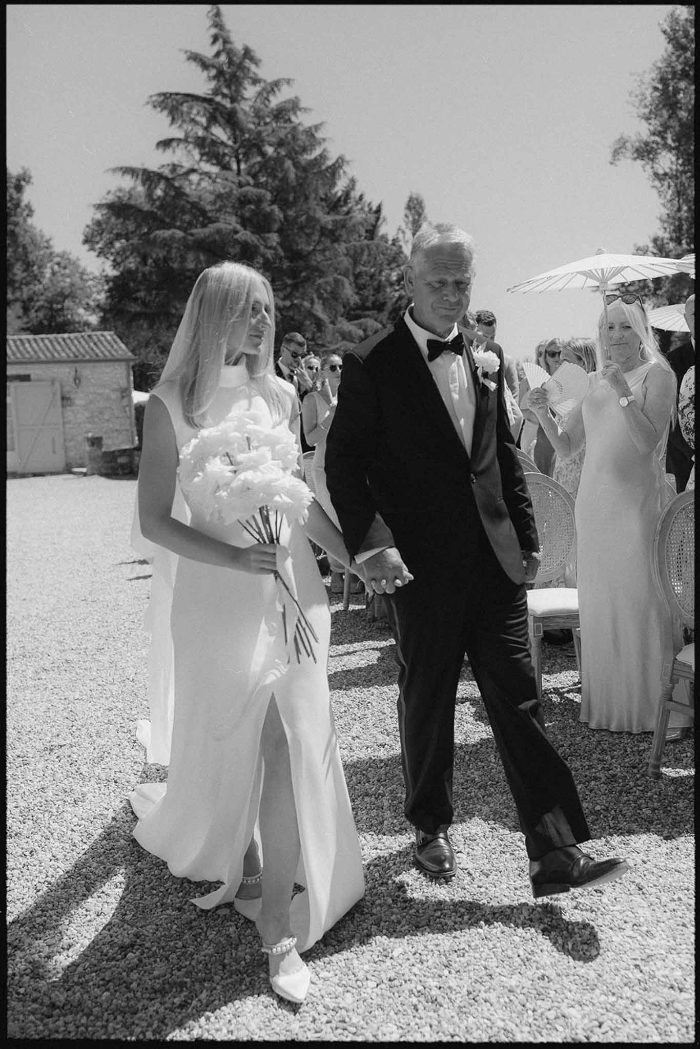 Black and white bride escorted down gravel aisle with bouquet veil and guests holding fans and parasols