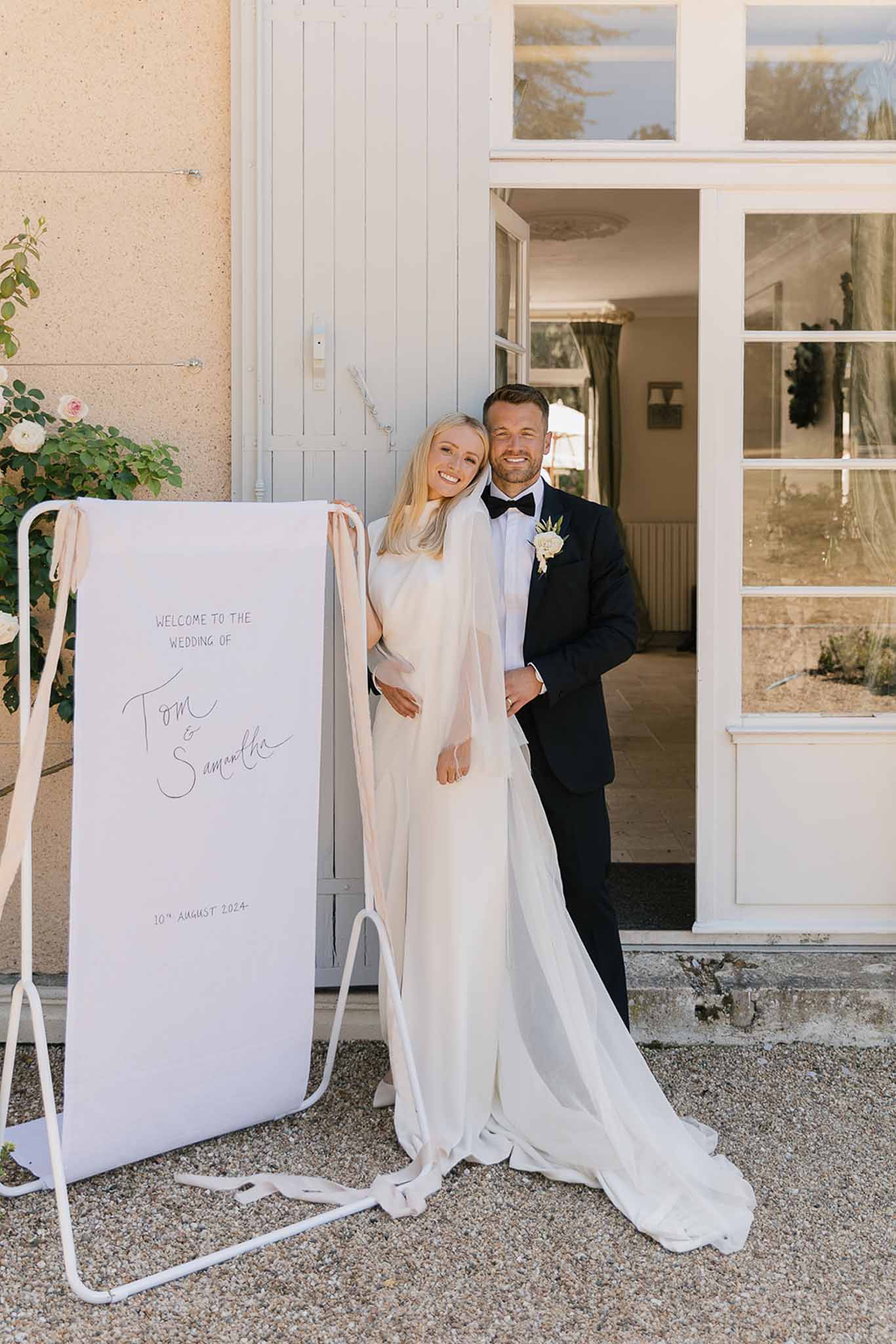 Bride in ivory satin slip gown and groom in black tuxedo beside calligraphy welcome sign at chateau entrance
