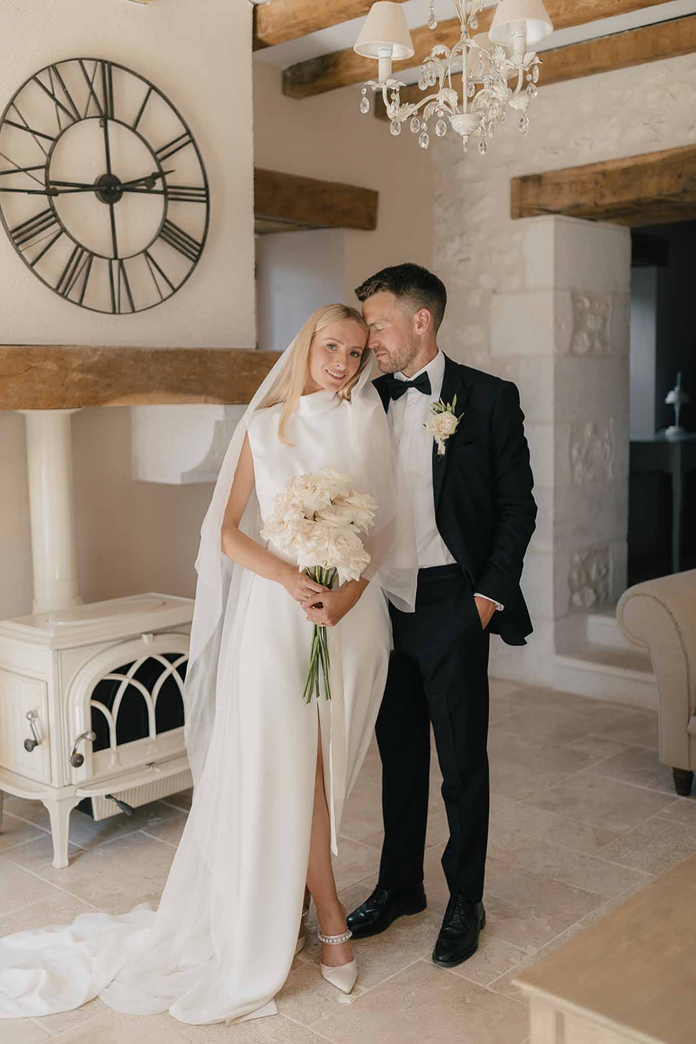 Bride in white satin column gown with cream peony bouquet and groom by limestone fireplace and chandelier
