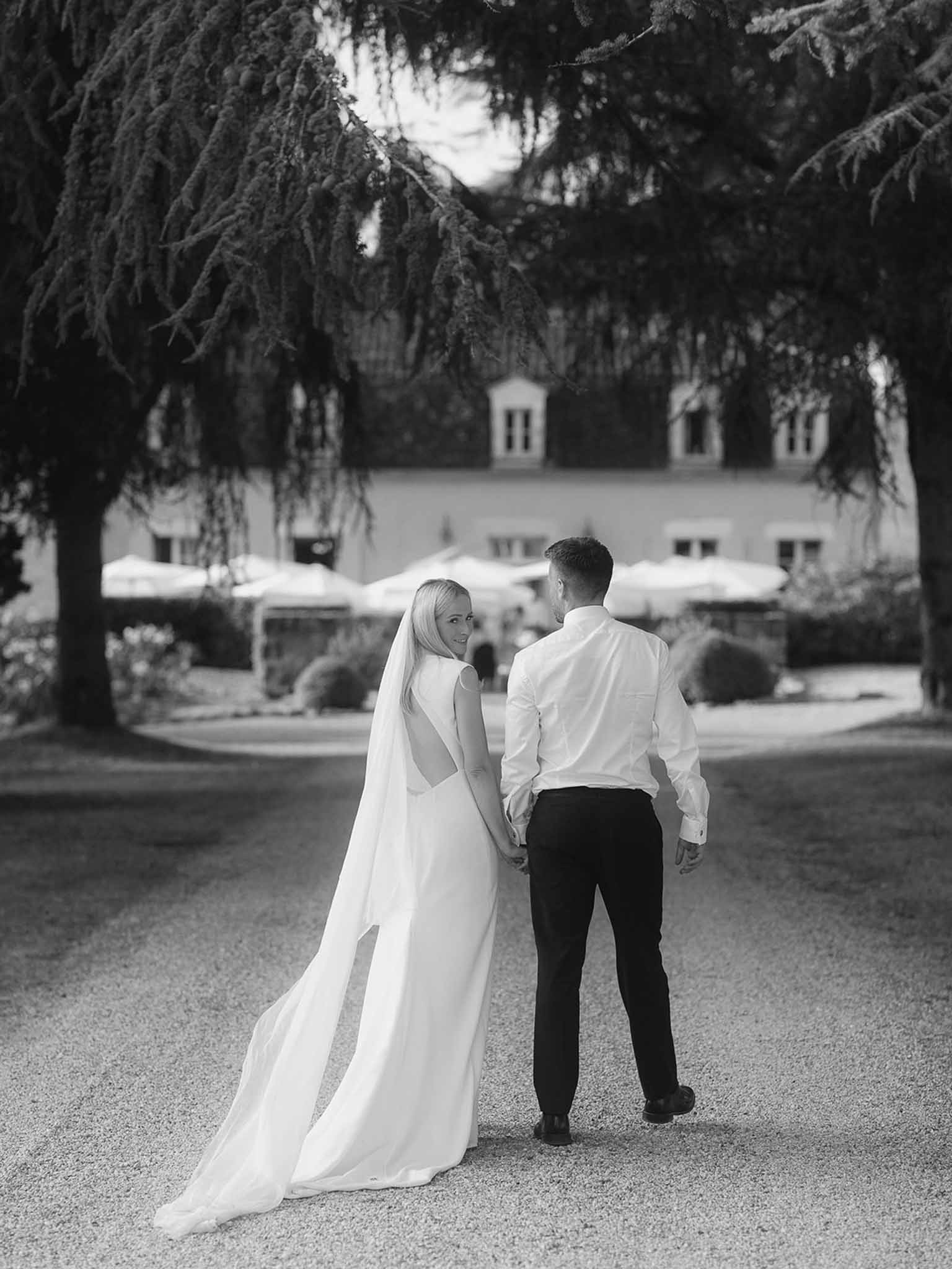 Black and white couple walking down tree-lined drive with cathedral veil trailing toward manor