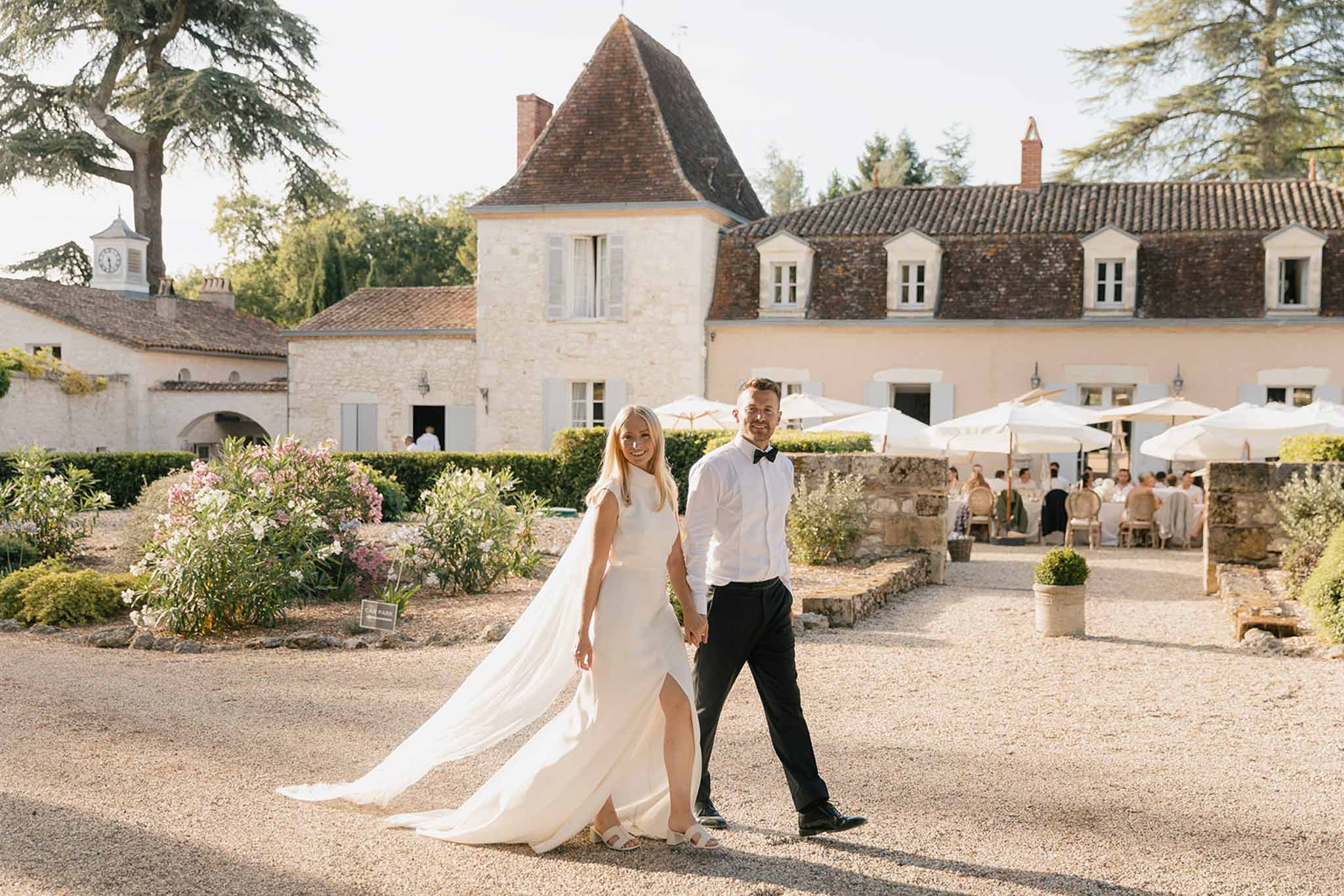 Couple holding hands walking toward camera bride in white cape-train gown guests under umbrellas and chateau tower behind