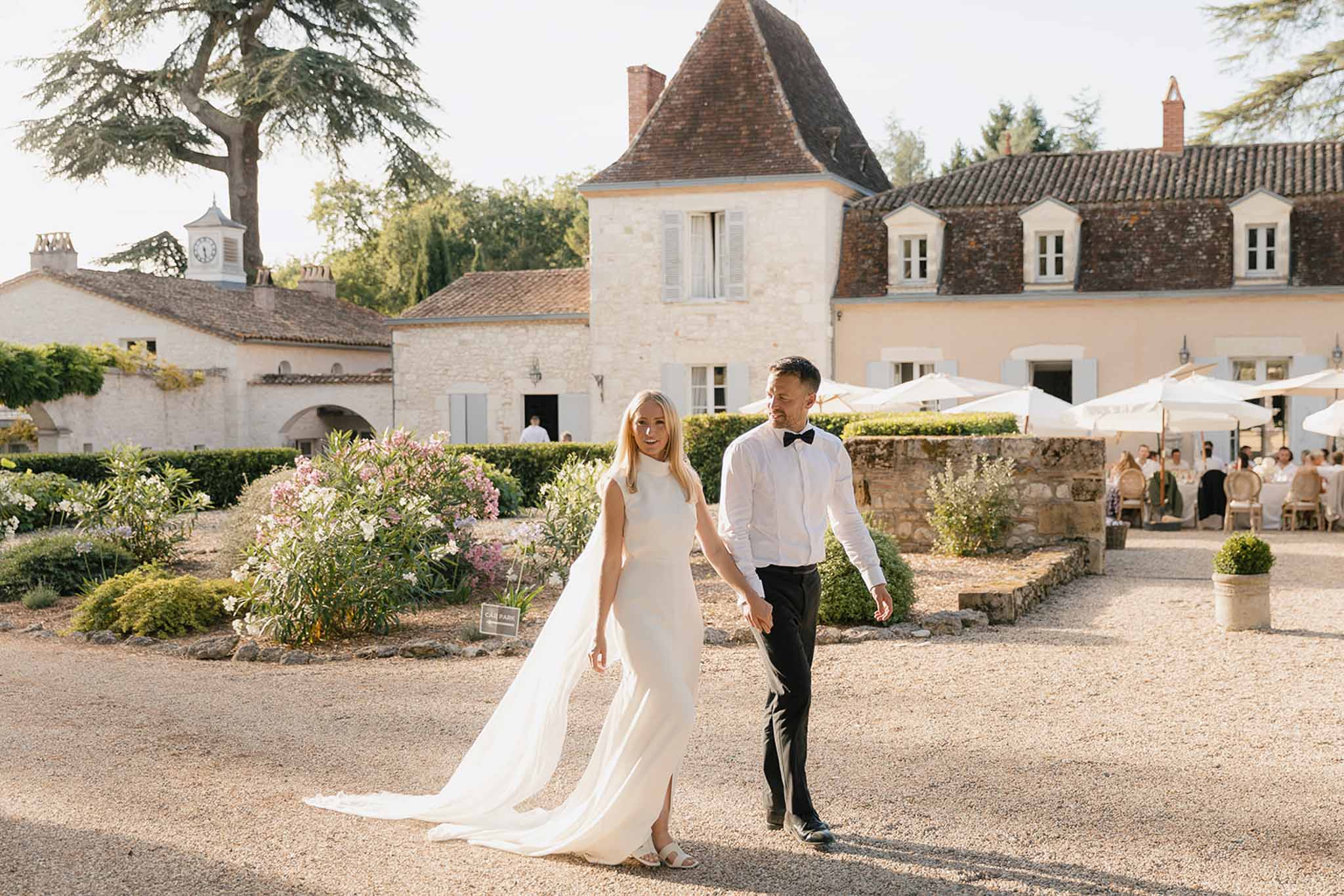 Bride in column gown with cape veil and groom walking through chateau courtyard with guest parasols