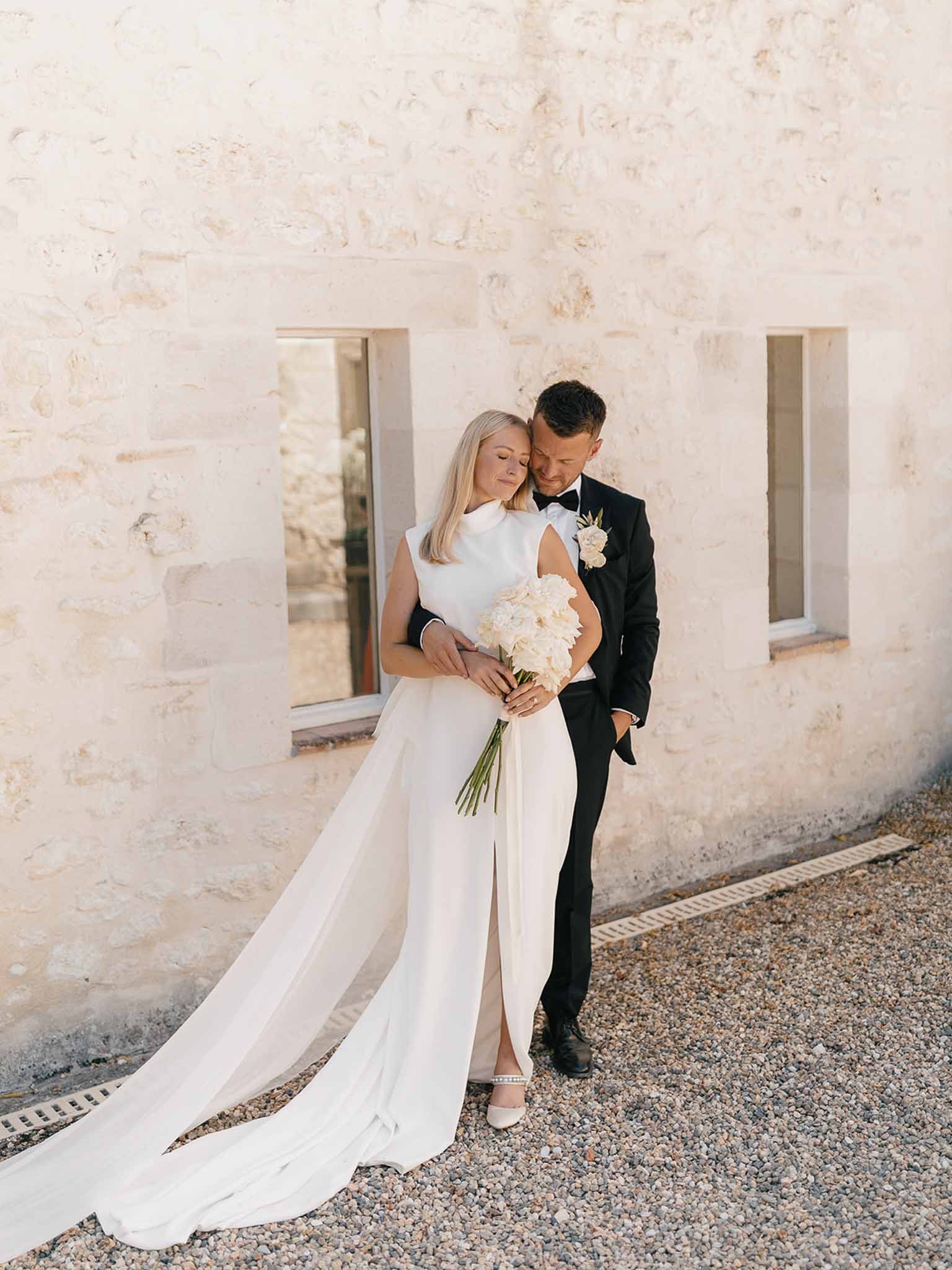 Groom embracing bride from behind against limestone wall, bride holding ivory dahlia bouquet