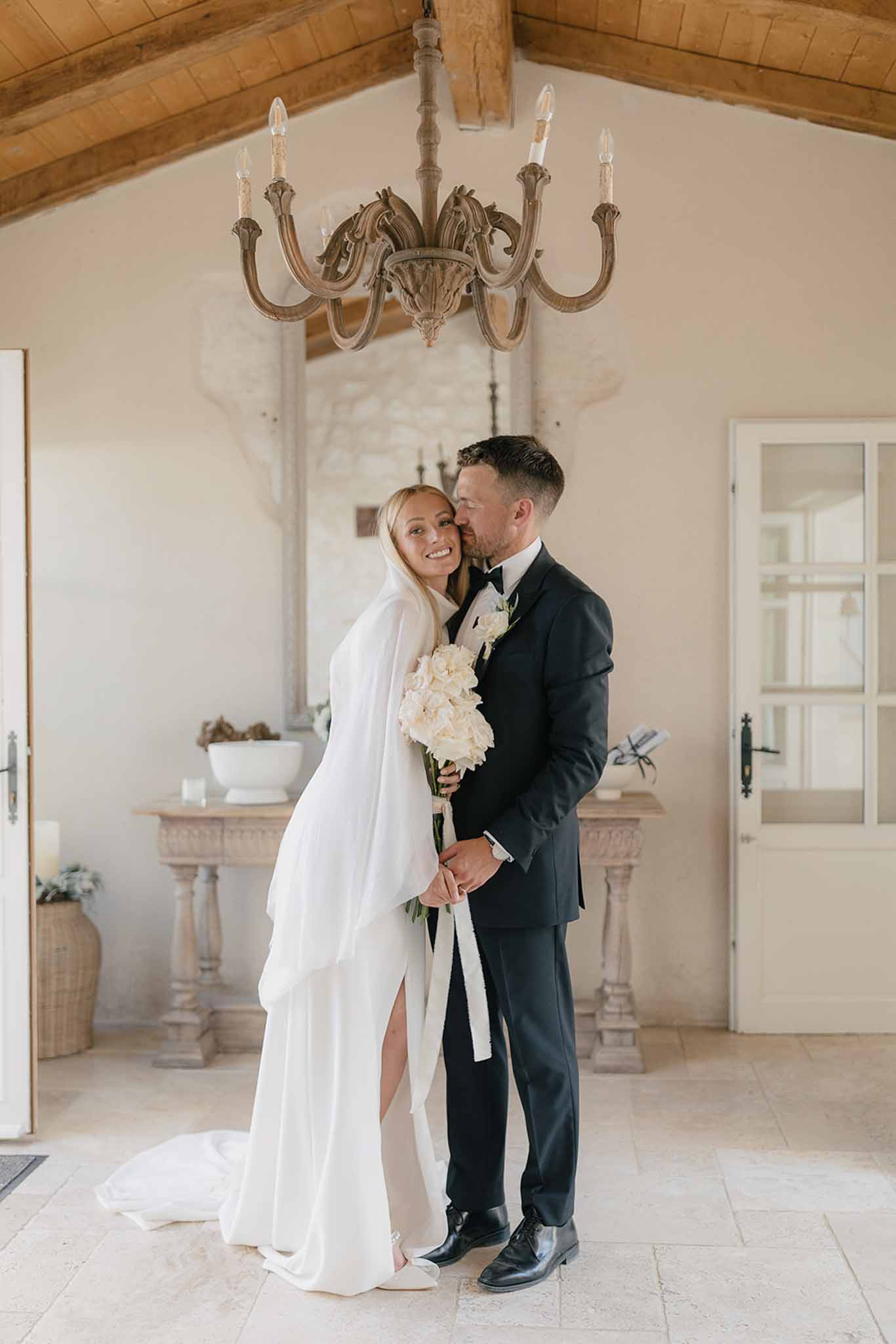 Groom kissing bride's cheek in whitewashed room with beam ceiling bride holding white hydrangea bouquet