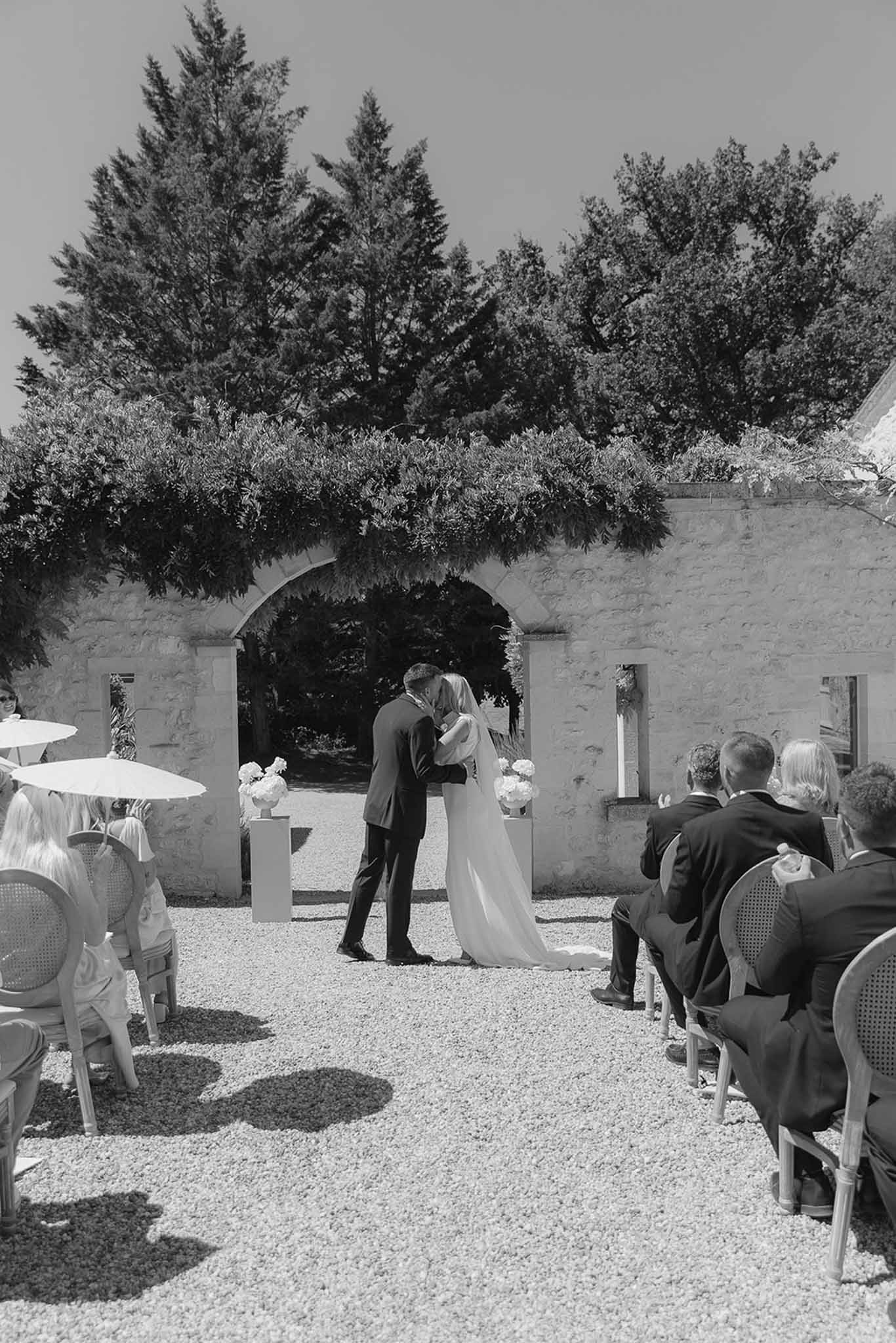Black and white wide shot of first kiss at outdoor ceremony under stone archway with seated guests and parasols