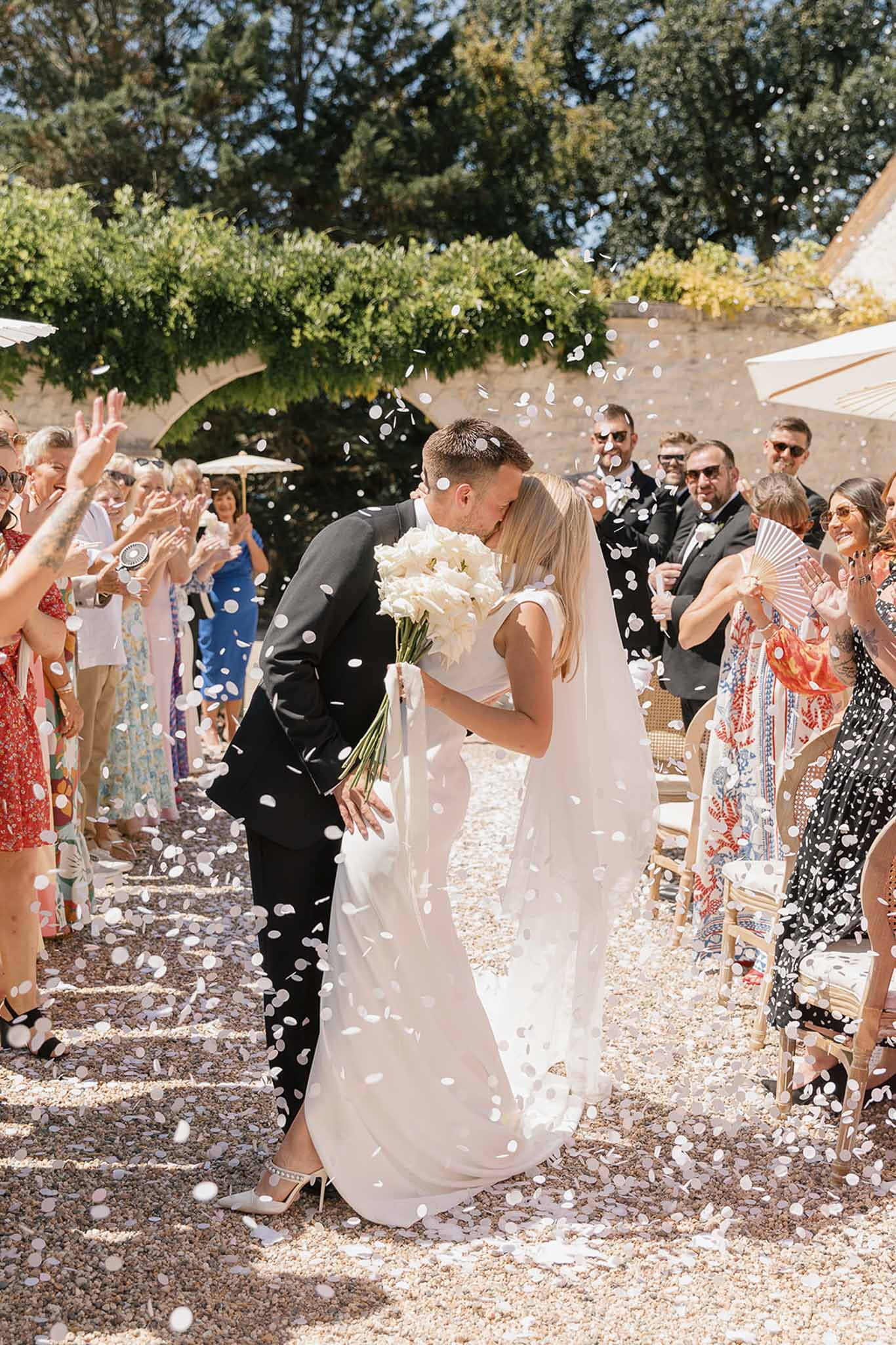 Bride and groom kiss during recessional as guests throw white confetti in a sunny stone courtyard